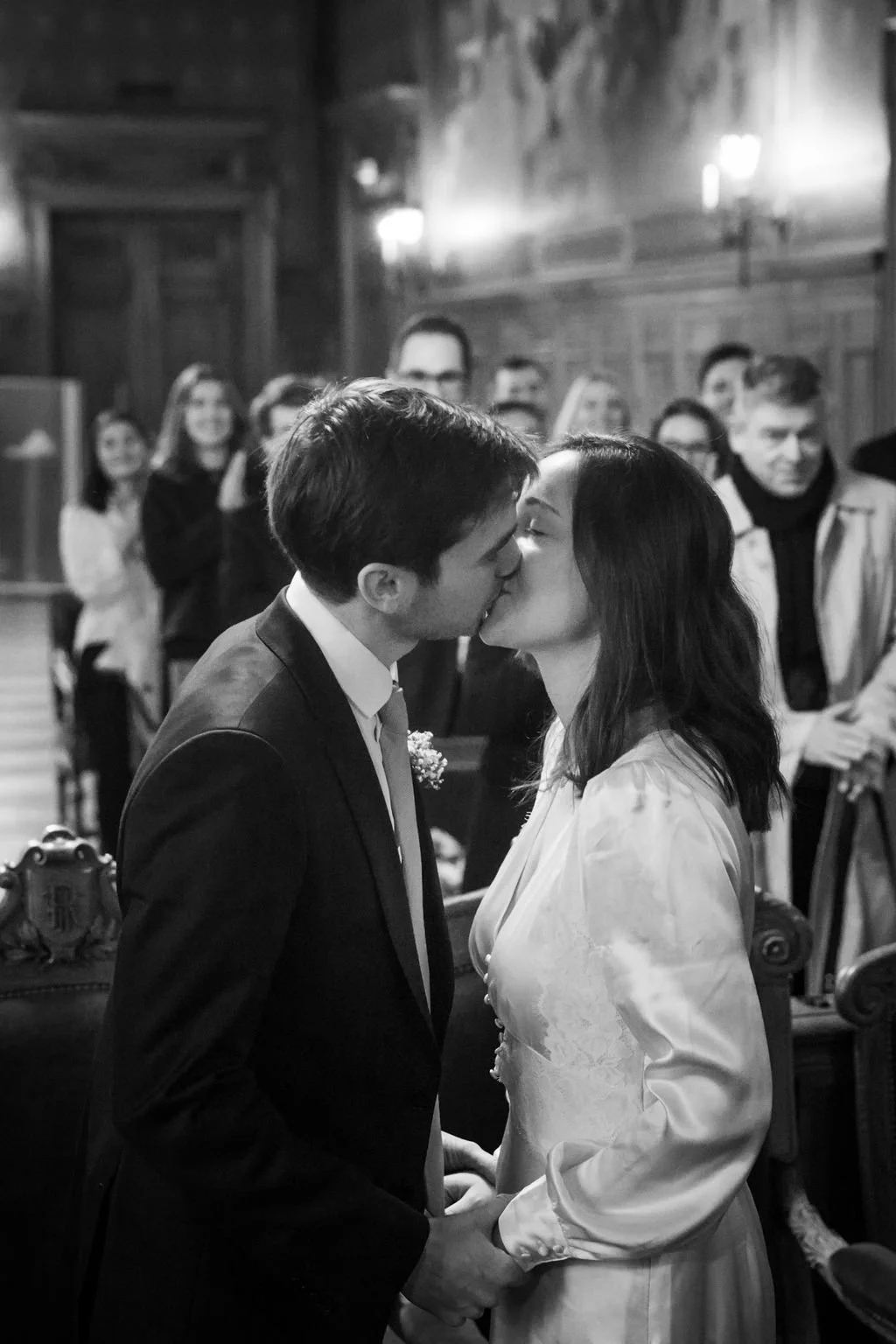 Un couple s'embrasse lors de leur mariage dans une salle avec des invités qui regardent et prennent des photos. Par votre photographe de mariage à paris