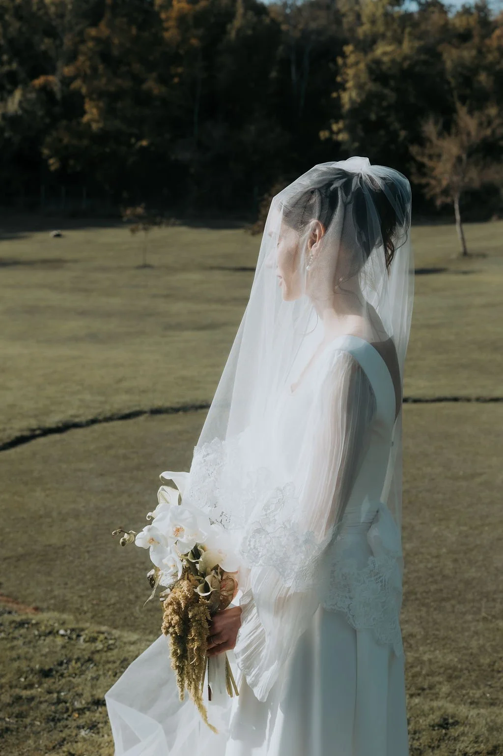 Femme en robe de mariée avec un voile, tenant un bouquet de fleurs blanches, en plein air avec un paysage de champs et arbres en arrière-plan. Par votre photographe de mariage à paris