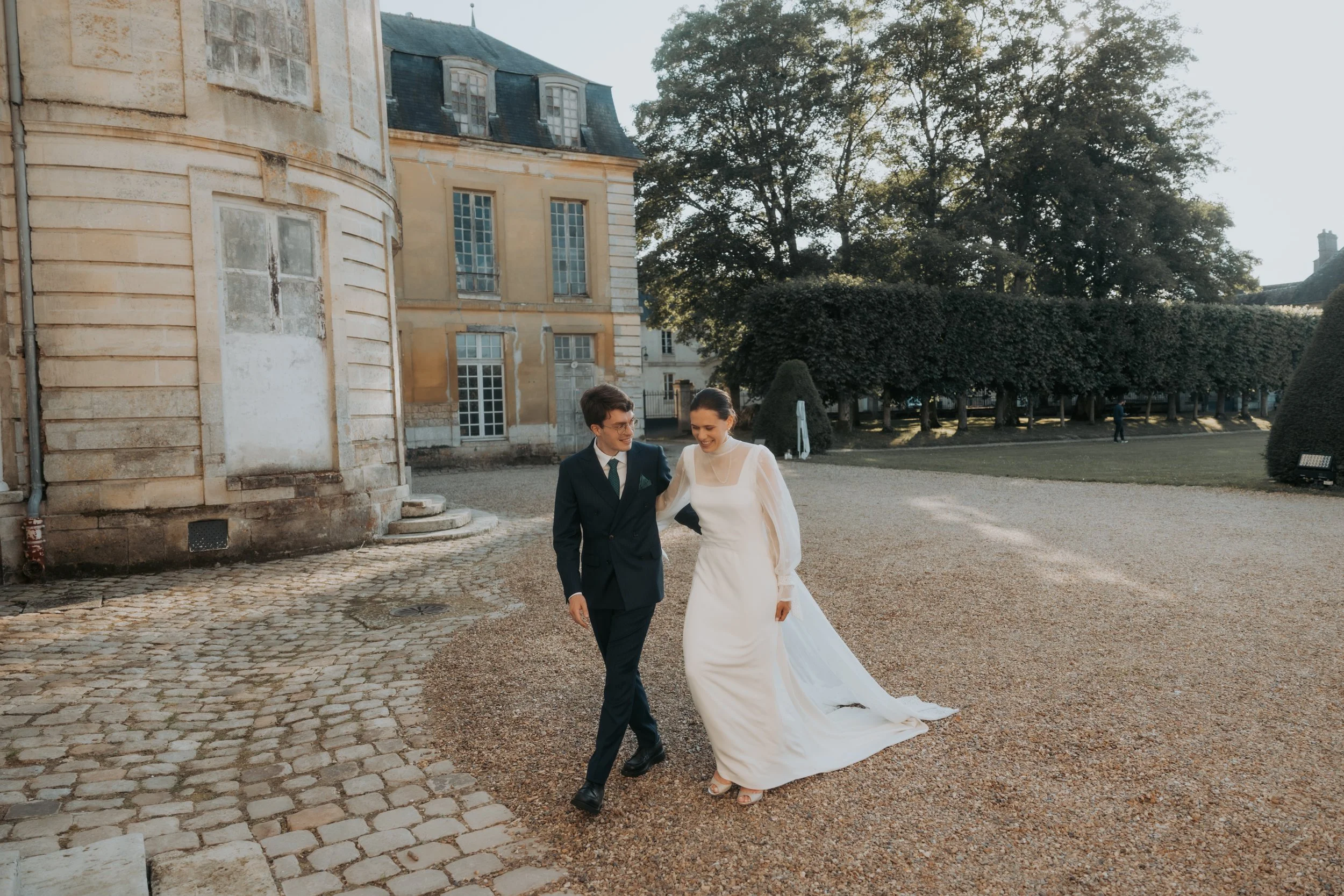 Un couple marié marche main dans la main devant un château en pierre avec un grand jardin, buissons et arbres en arrière-plan, lors d'une journée ensoleillée. Le jour de leur mariage en chateau à paris