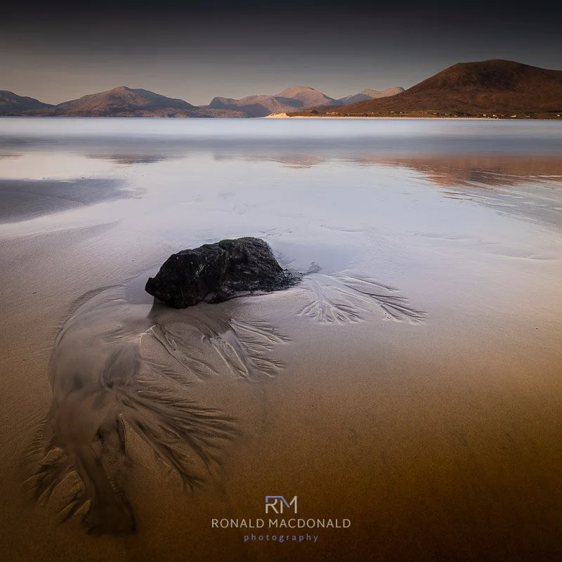 A calm Harris beach with a large black rock in the foreground, wet sand patterns around it, and distant mountains under a cloudy sky.