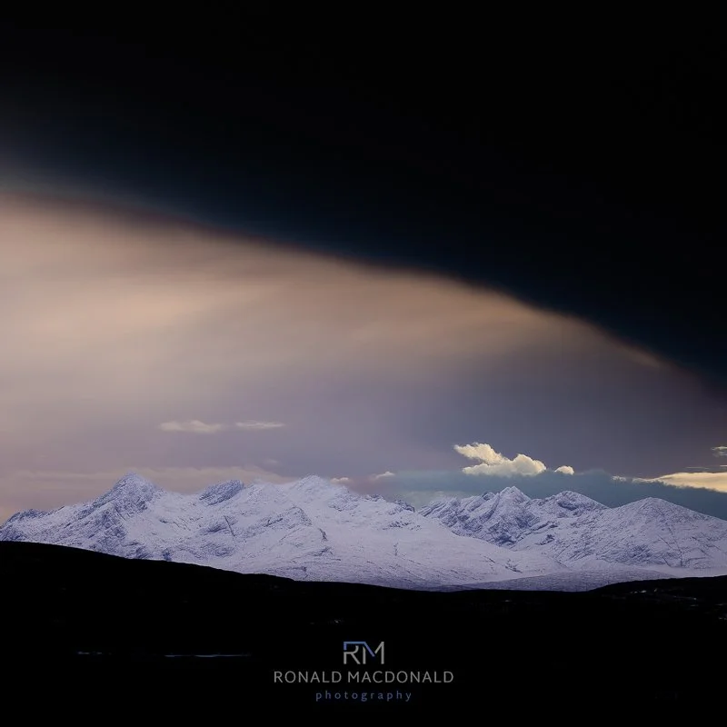 Snowy Cuillin peaks under a dramatic dark sky with soft light breaking through, photographed by Ronald MacDonald.