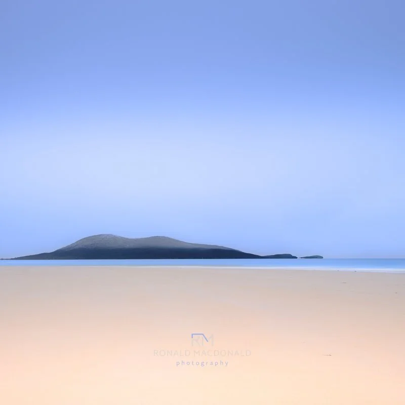 A solitary figure walks along a beach, with a distant mountain range visible against the horizon.