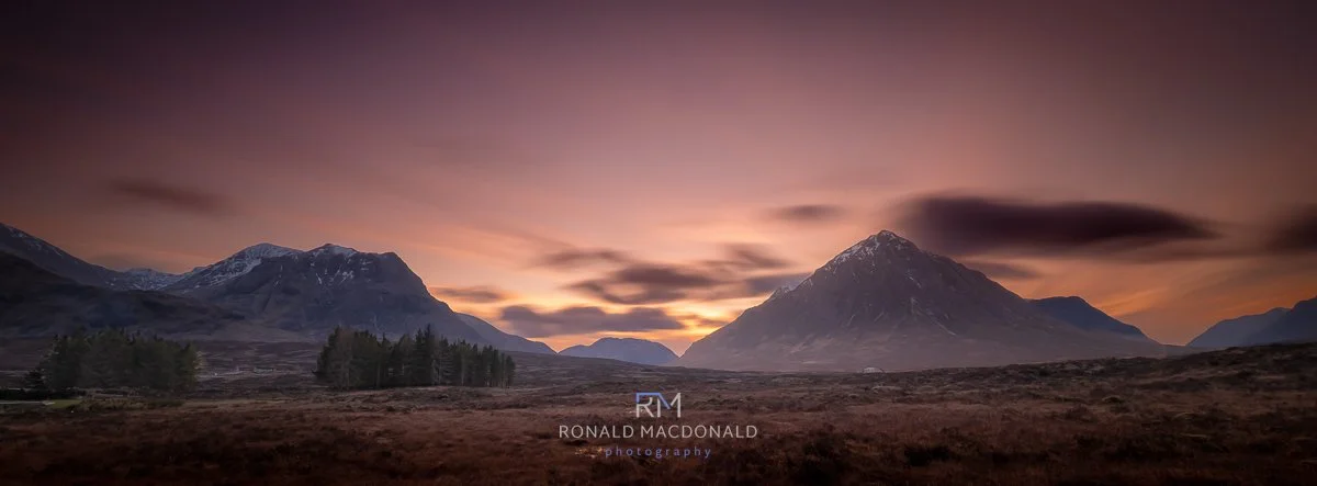 Glencoe mountain landscape at sunset with purple and orange sky, snow-capped peaks, and forested area in foreground.