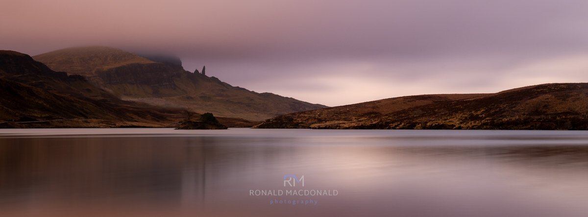 The pink huues of pre sunset refelct over Loch Fada Skye  with a long cloud above the old man of Storr