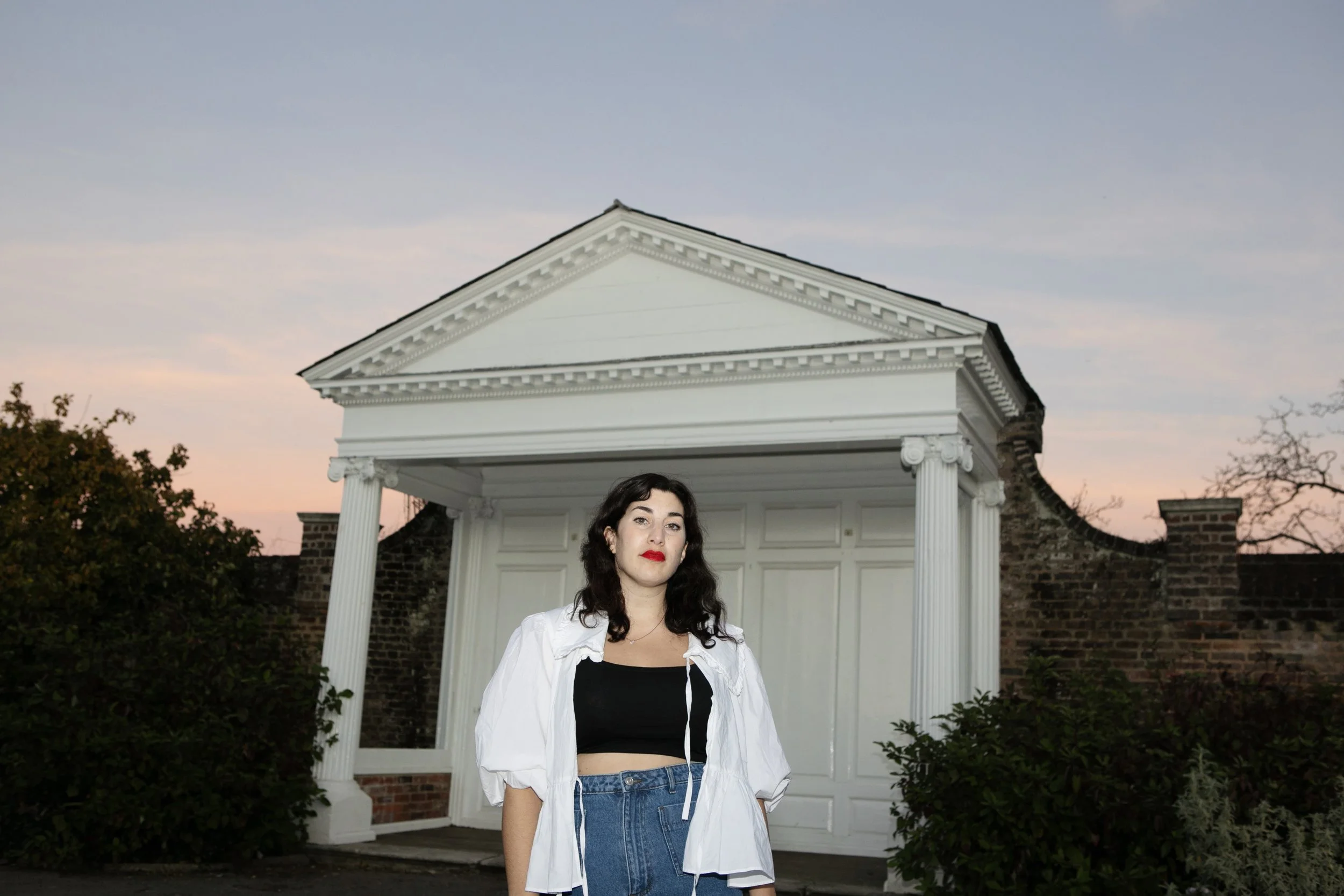 A woman with dark, wavy hair and red lipstick standing in front of a white, classical-style building with columns at sunset.
