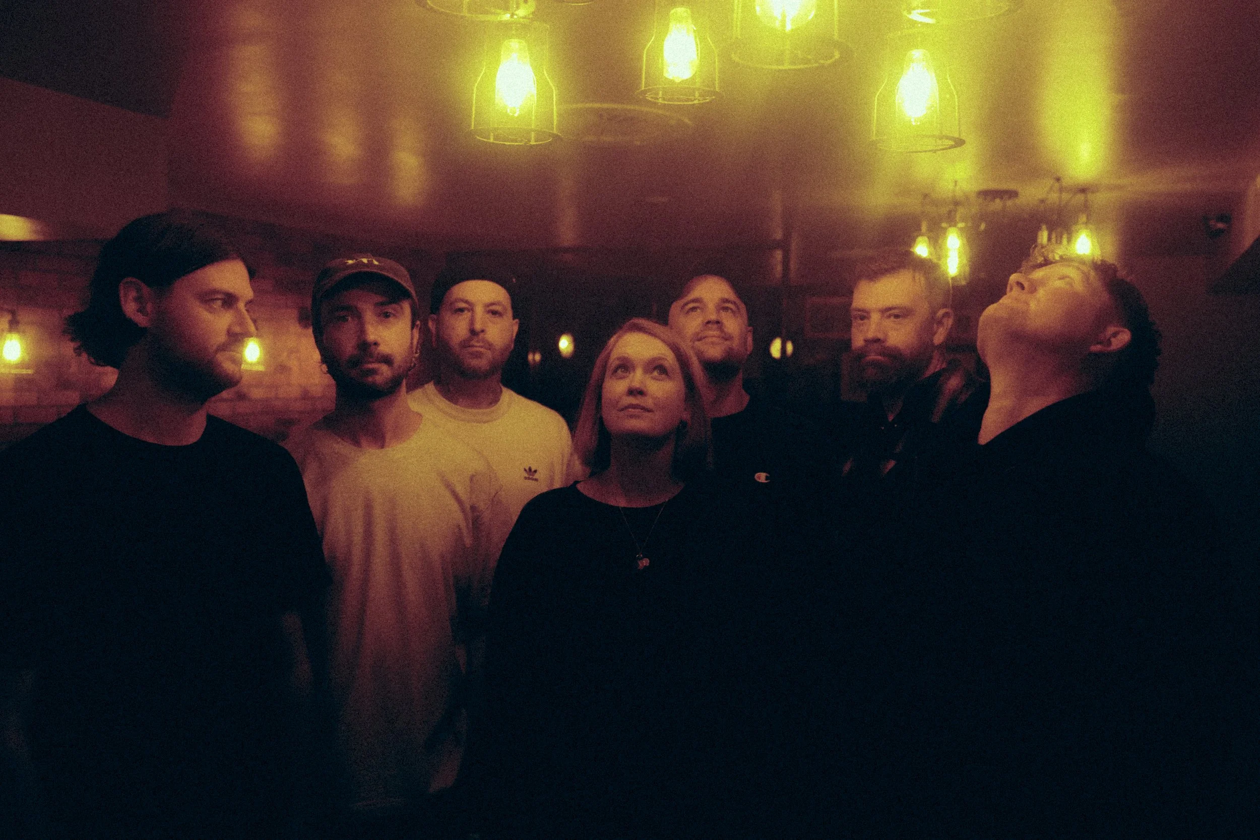 A group of seven people standing together indoors, some with serious expressions, against a plain light-colored wall with some plants in the corner.