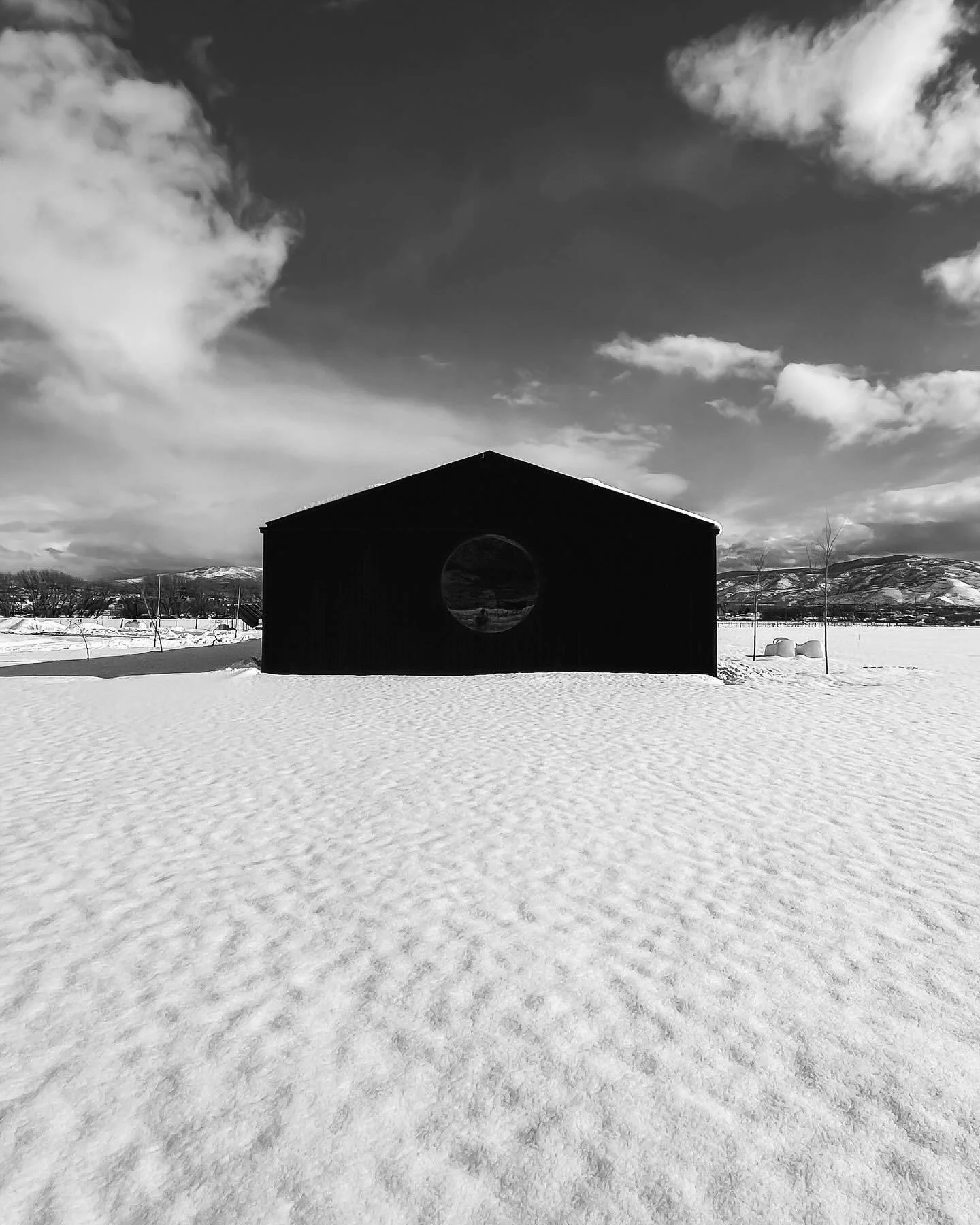 A black barn with a circular window, set in a snowy landscape under a cloudy sky with mountains in the background.