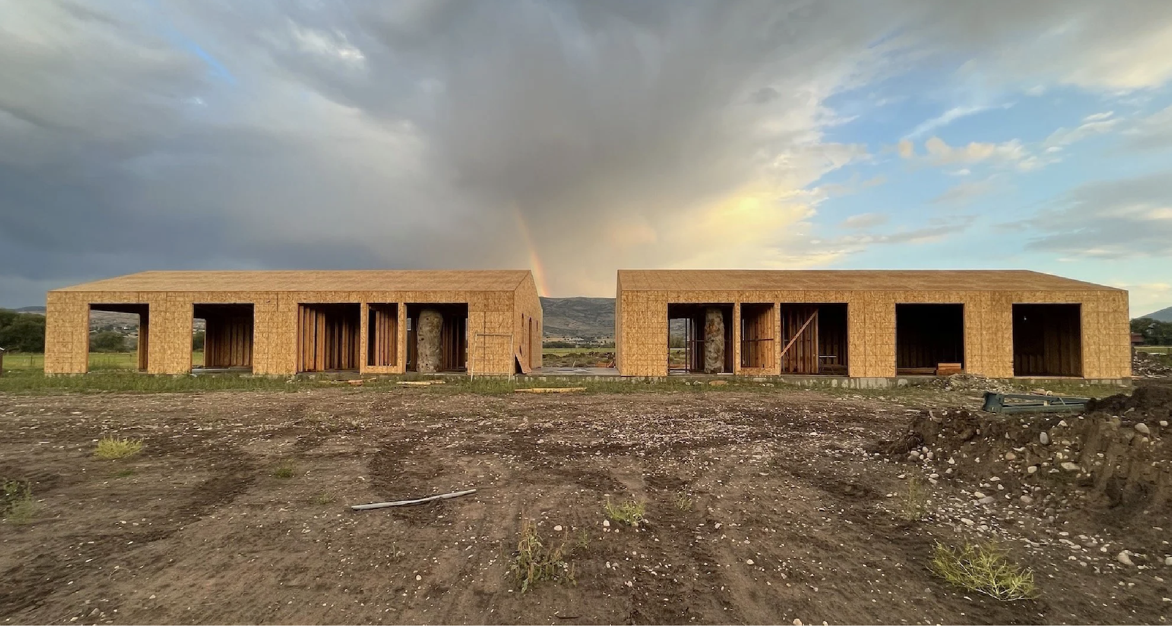 Two wooden house frames under construction with a dirt ground in foreground and a cloudy sky with a rainbow in the background.