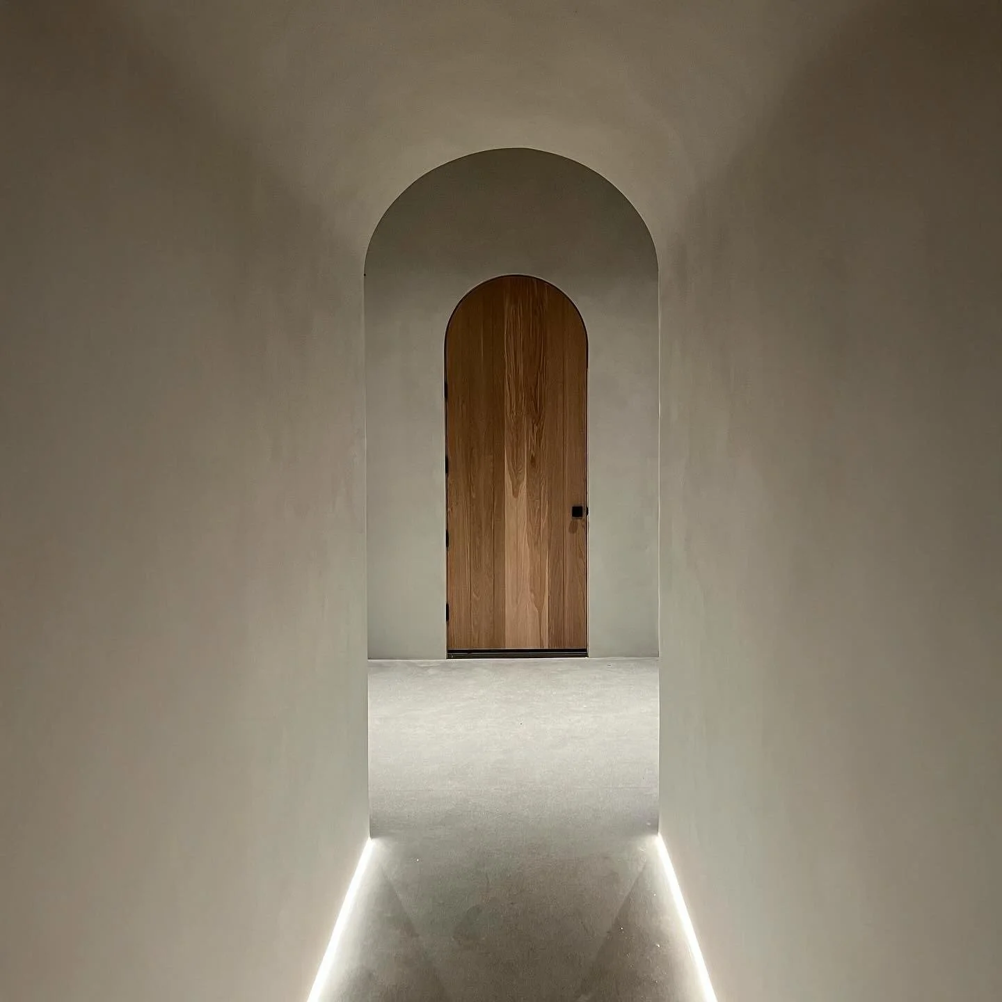 Minimalist hallway with soft lighting along the baseboards leads to a closed wooden door within an arched doorway.