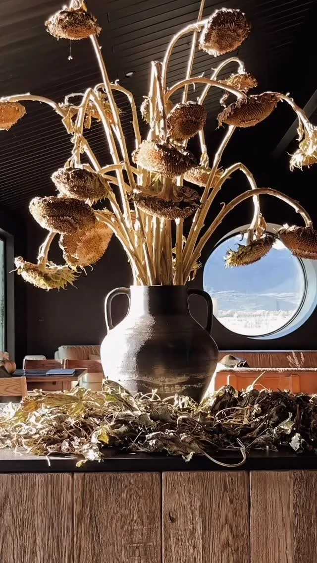 A large black vase with sunflower stems and dried sunflower heads on a table, with some dried leaves and stems around it. In the background, a window shows a cloudy sky.