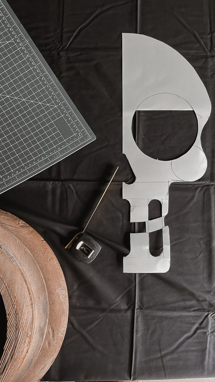 A black tablecloth with woodworking tools and a paper template. The template is cutout in the shape of a skull with a large circular hole in the forehead area, possibly for a costume or decoration. There is a large spool of pinkish-brown thread or tw