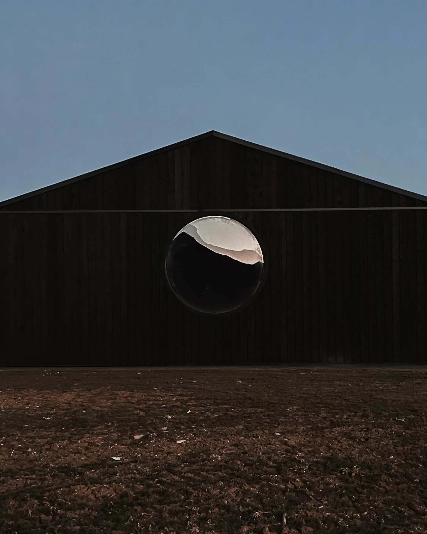A dark barn with a circular window that reflects mountains and the sky, set against an evening sky.