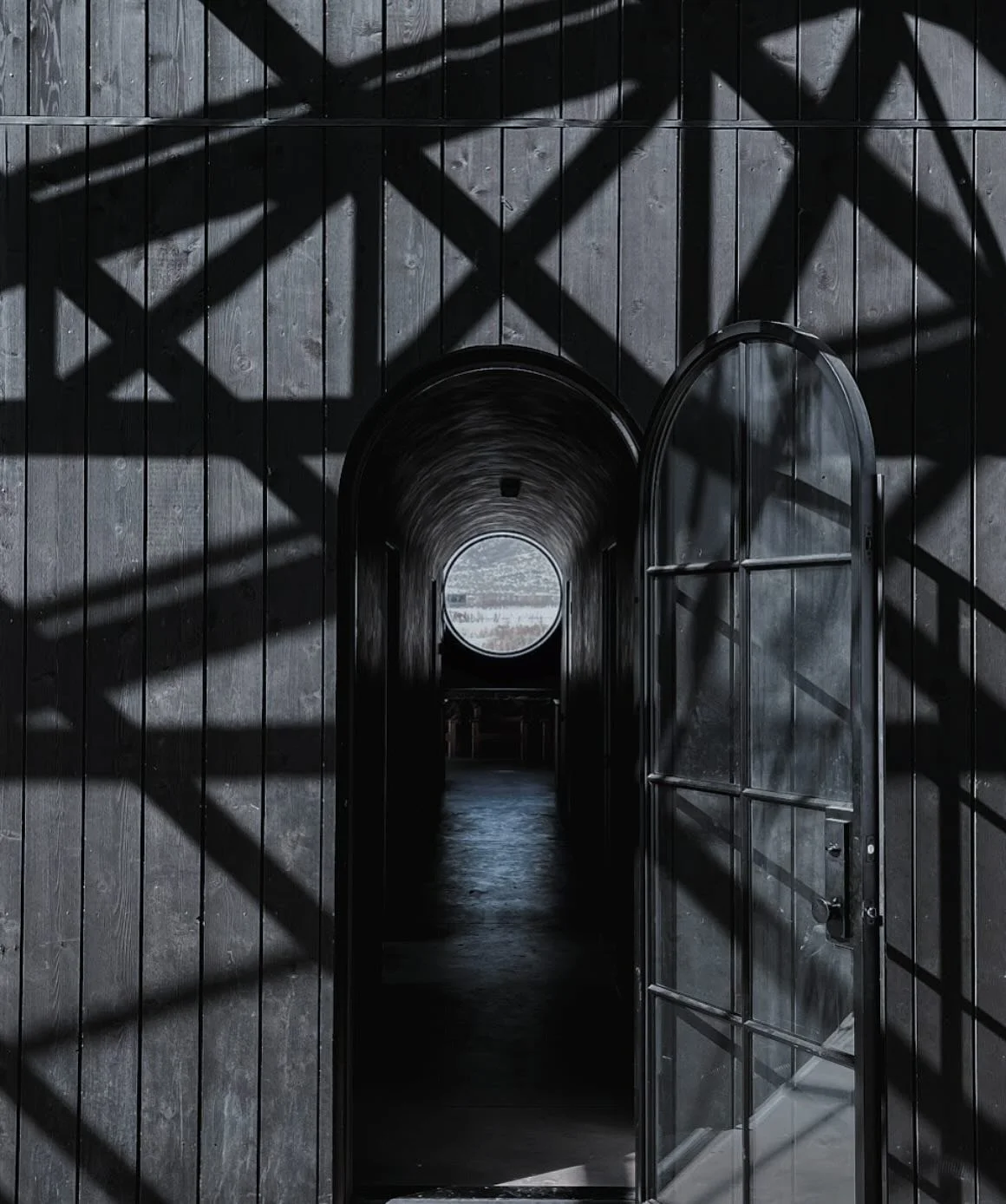 Open glass door leading to dark tunnel with a circular opening at the end, shadows cast on wooden wall outside.