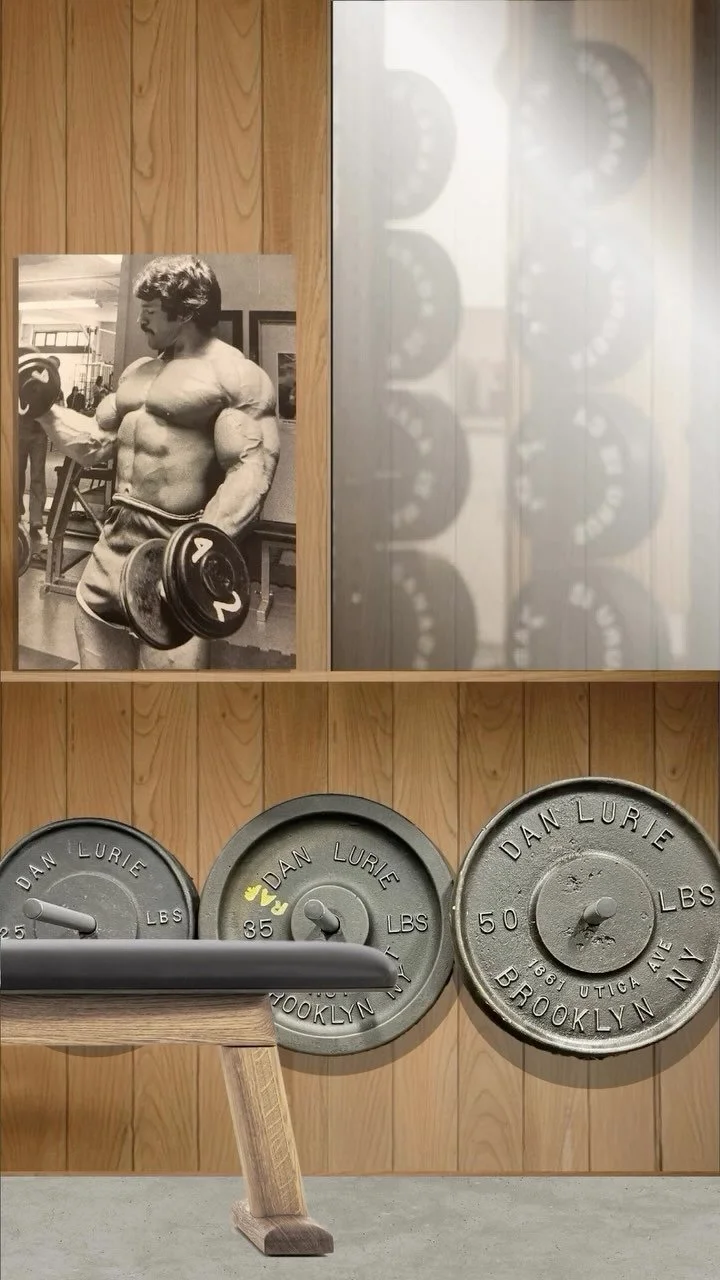Black and white poster of a muscular young man with curly hair lifting a dumbbell in a gym setting, with a wooden wall background. Below, three vintage metal weight plates labeled "Dan Lurie" in different sizes are leaning against a wooden wall, with