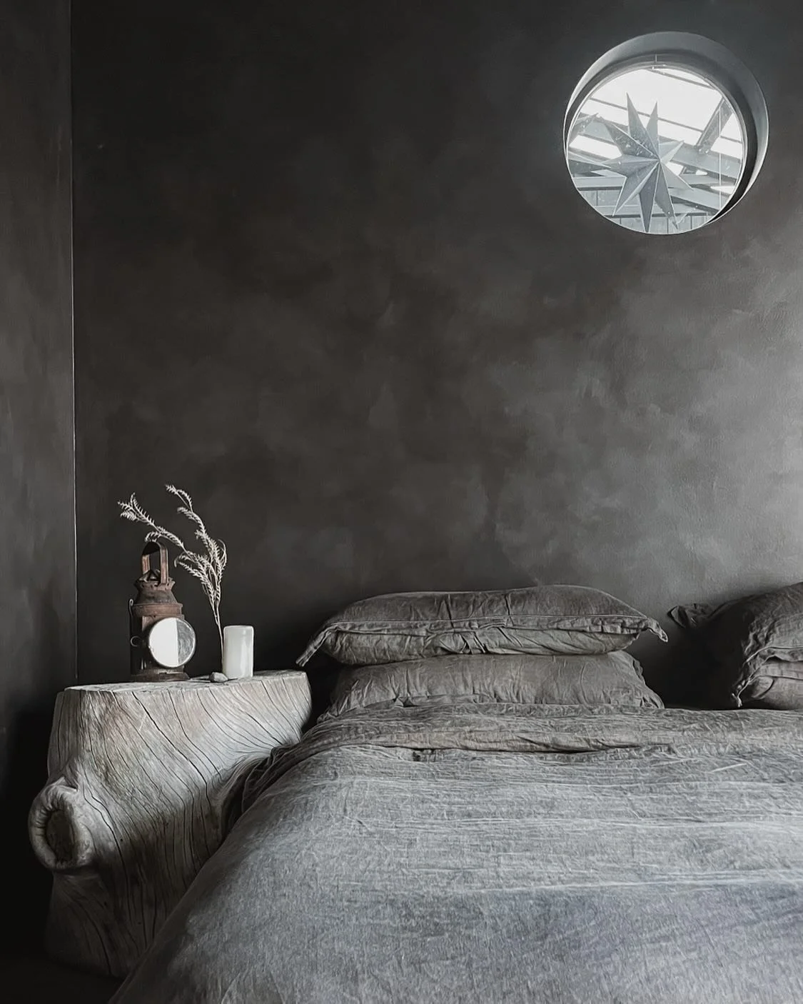 A bedroom with dark walls, a round window looking outside at a star-shaped structure, a bed with gray bedding, and a wooden nightstand with decor including a lantern, a candle, and dried plants.