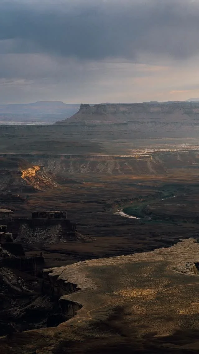 A vast canyon landscape under cloudy sky, with layered rock formations, a river flowing through the valley, and distant plateau edges.