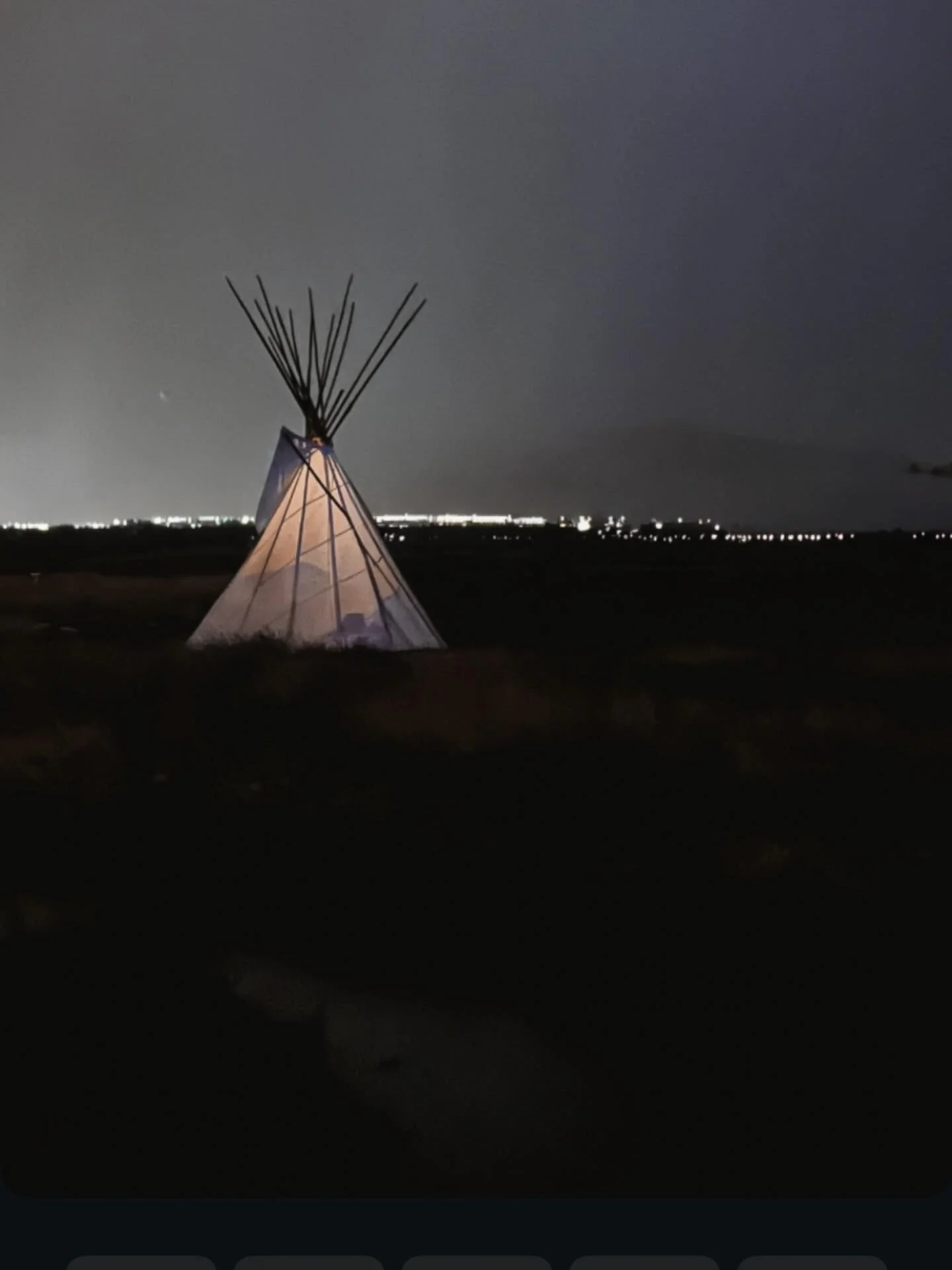 Nighttime scene featuring a traditional teepee structure illuminated from within, set against a dark sky and distant city lights on the horizon.