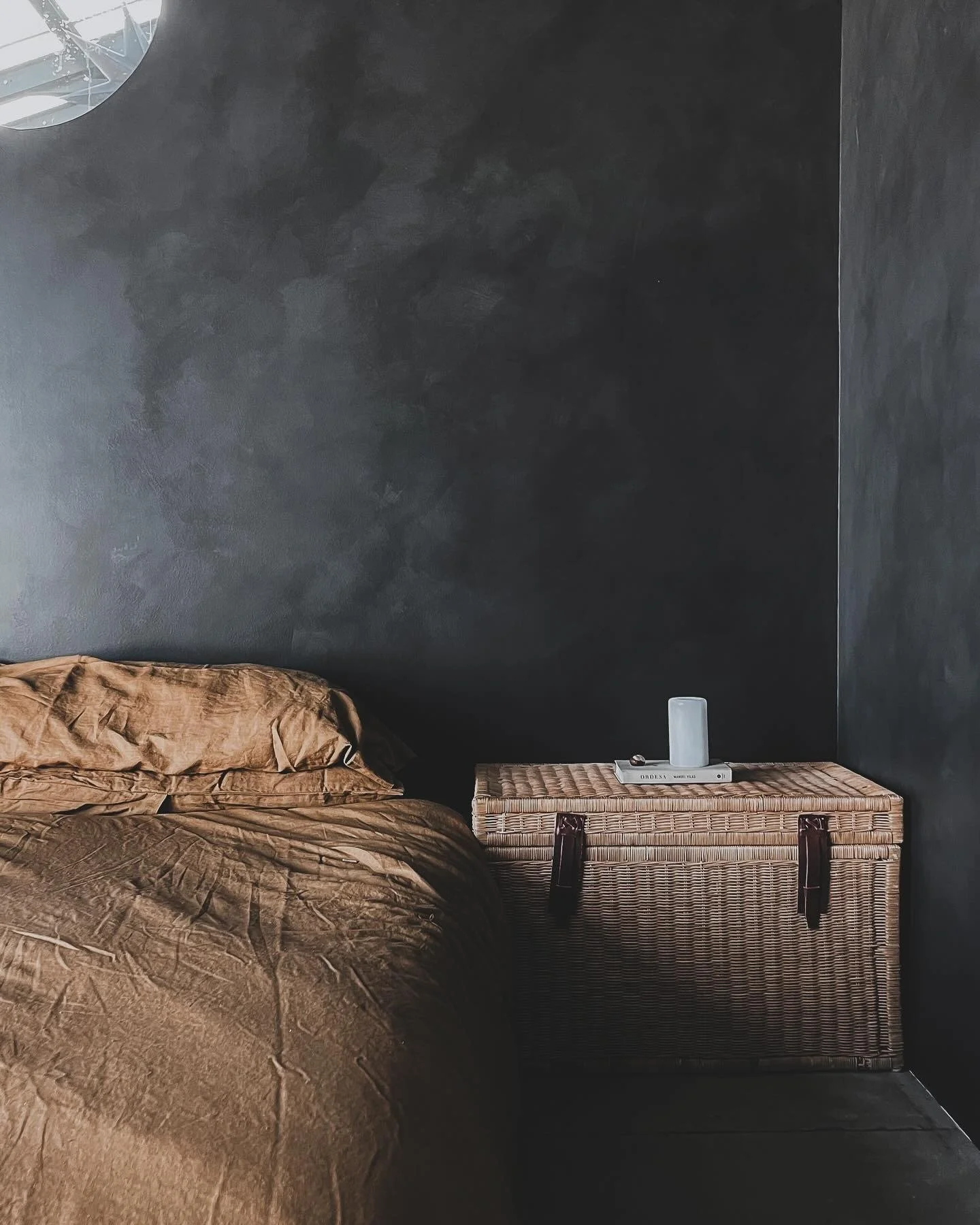 A bedroom with a dark textured wall, a brown bed with wrinkled bedding, and a wicker nightstand with a white book and a white decorative object on top.