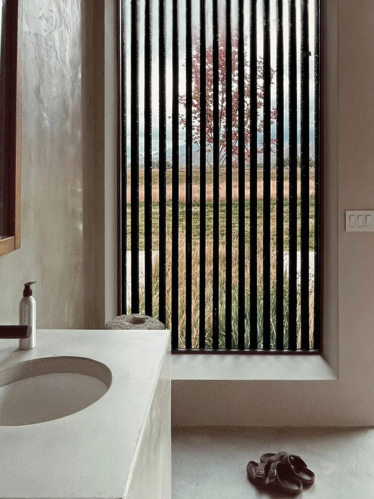A bathroom with a white countertop and a sink, a bottle of soap, a pair of black sandals on the floor, and a large window with black vertical slats showing an outdoor scene with a tree and blue sky.