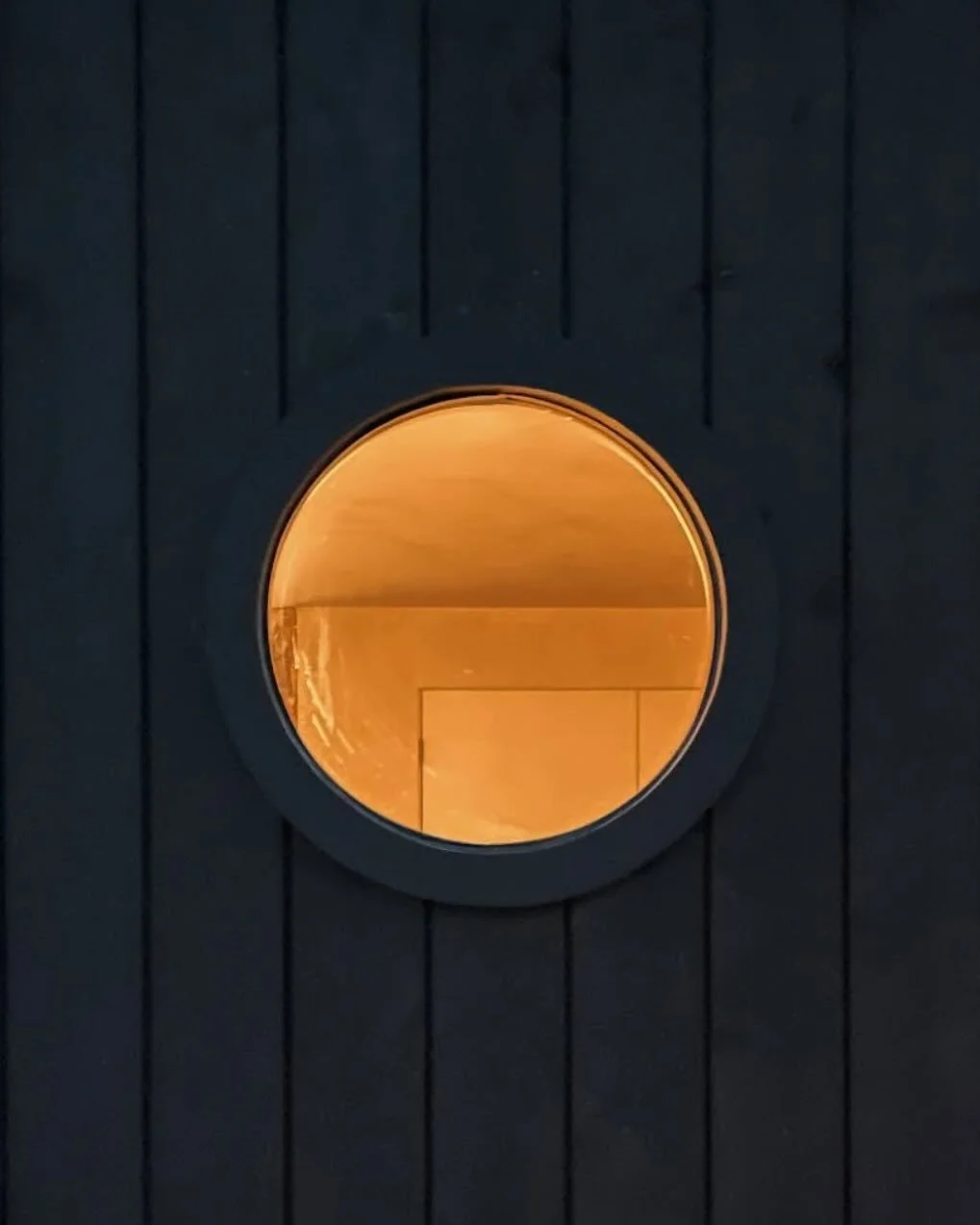Round window with warm yellow light, set into a black wooden wall with vertical planks.