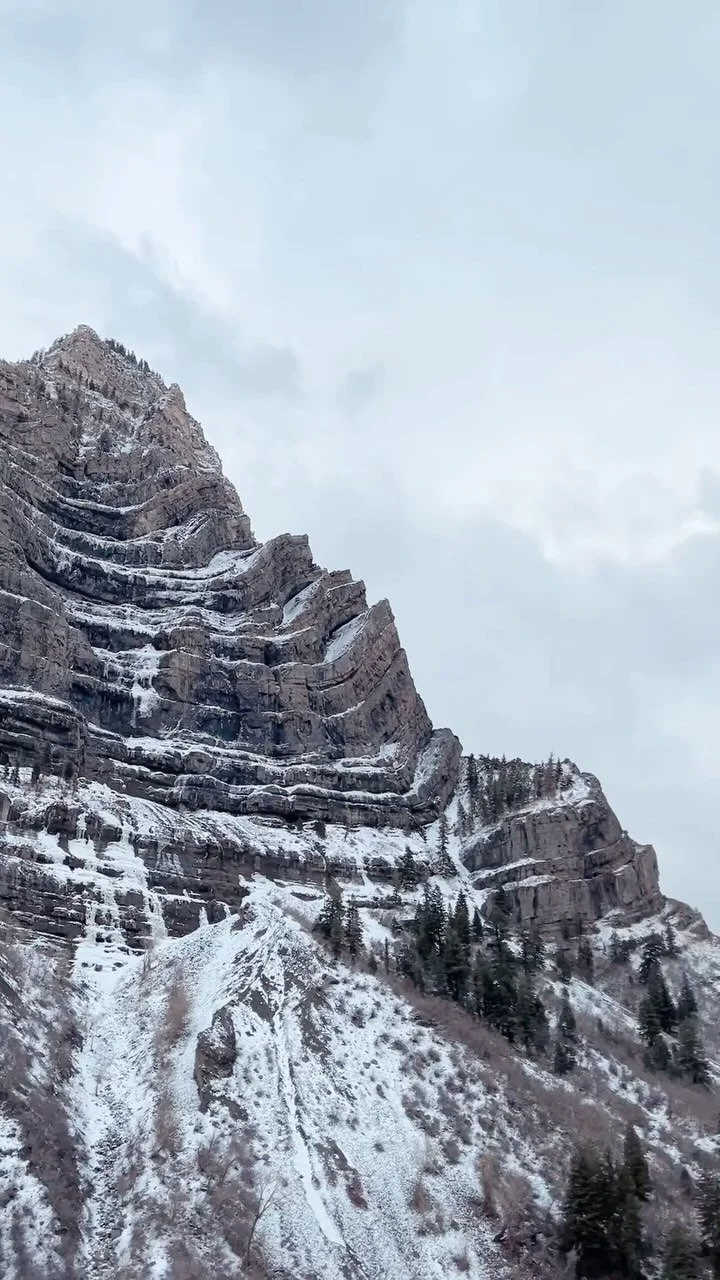 Snow-dusted rugged mountain peak with layered rock formations and sparse trees, under a cloudy sky.