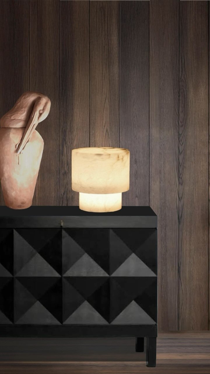 Decorative interior scene with a pink pelican sculpture, a glowing beige table lamp, and a black geometric cabinet against a dark wood-paneled wall.