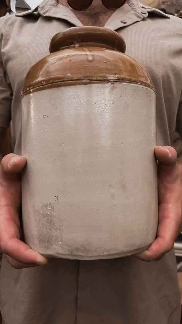 Person holding a large ceramic or pottery jar with a rounded lid, beige body, and brown top.
