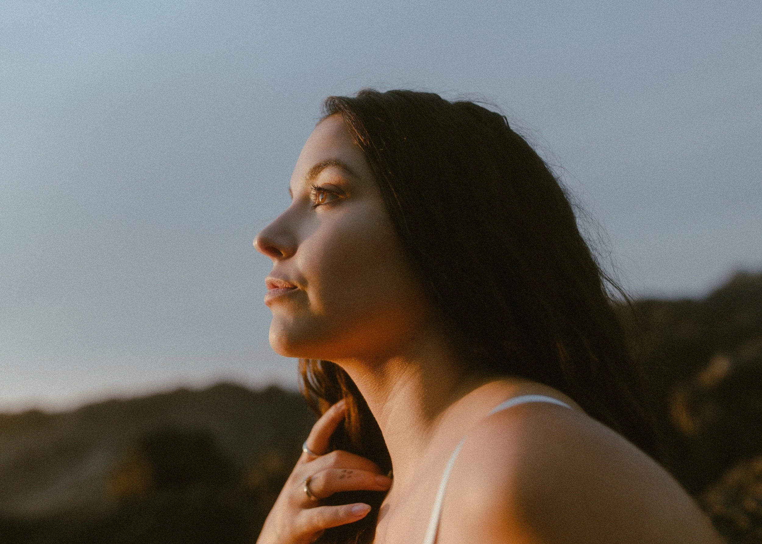 Close-up side view of brunette with flowing hair massaging her jawline outdoors at dusk, illustrating at home facial massage tutorial.