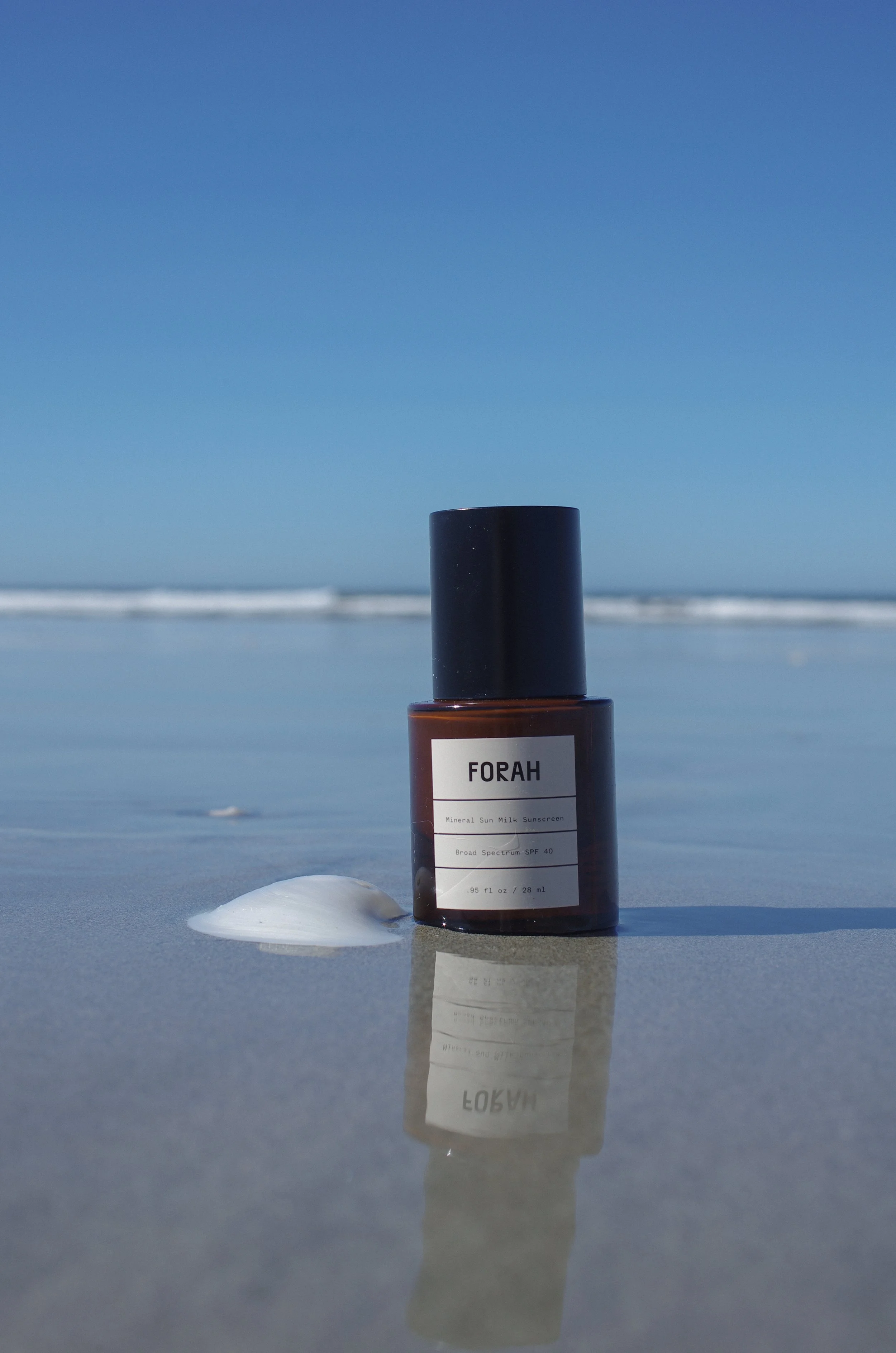 Bottle of Forah Mineral Sun Milk Sunscreen SPF40 on wet beach sand, reflecting in shallow ocean water with blue sky and waves.