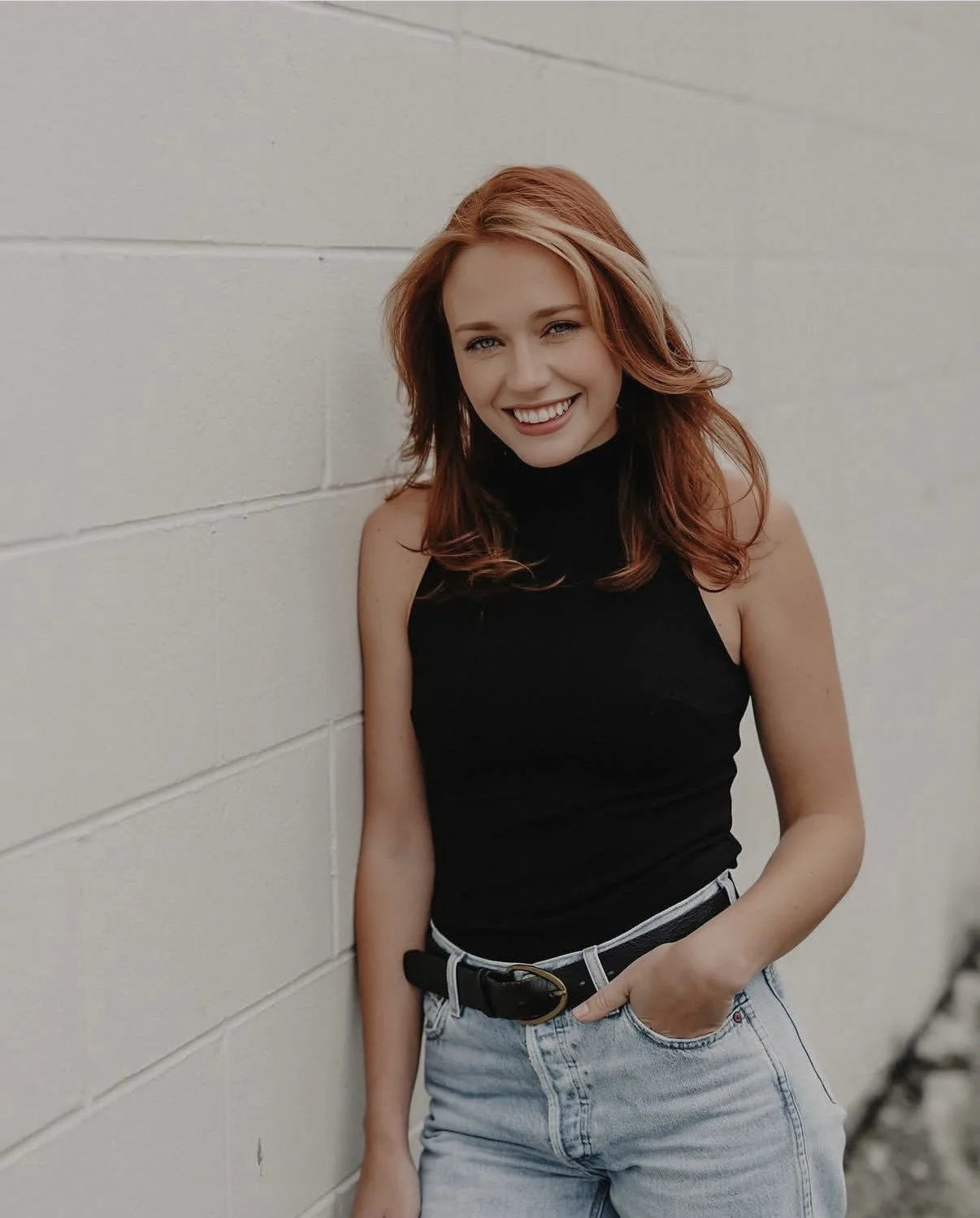 Teegan Tooth Whitening & Tooth Gem technician smiles confidently against a white brick wall, wearing a black turtleneck tank and light jeans.