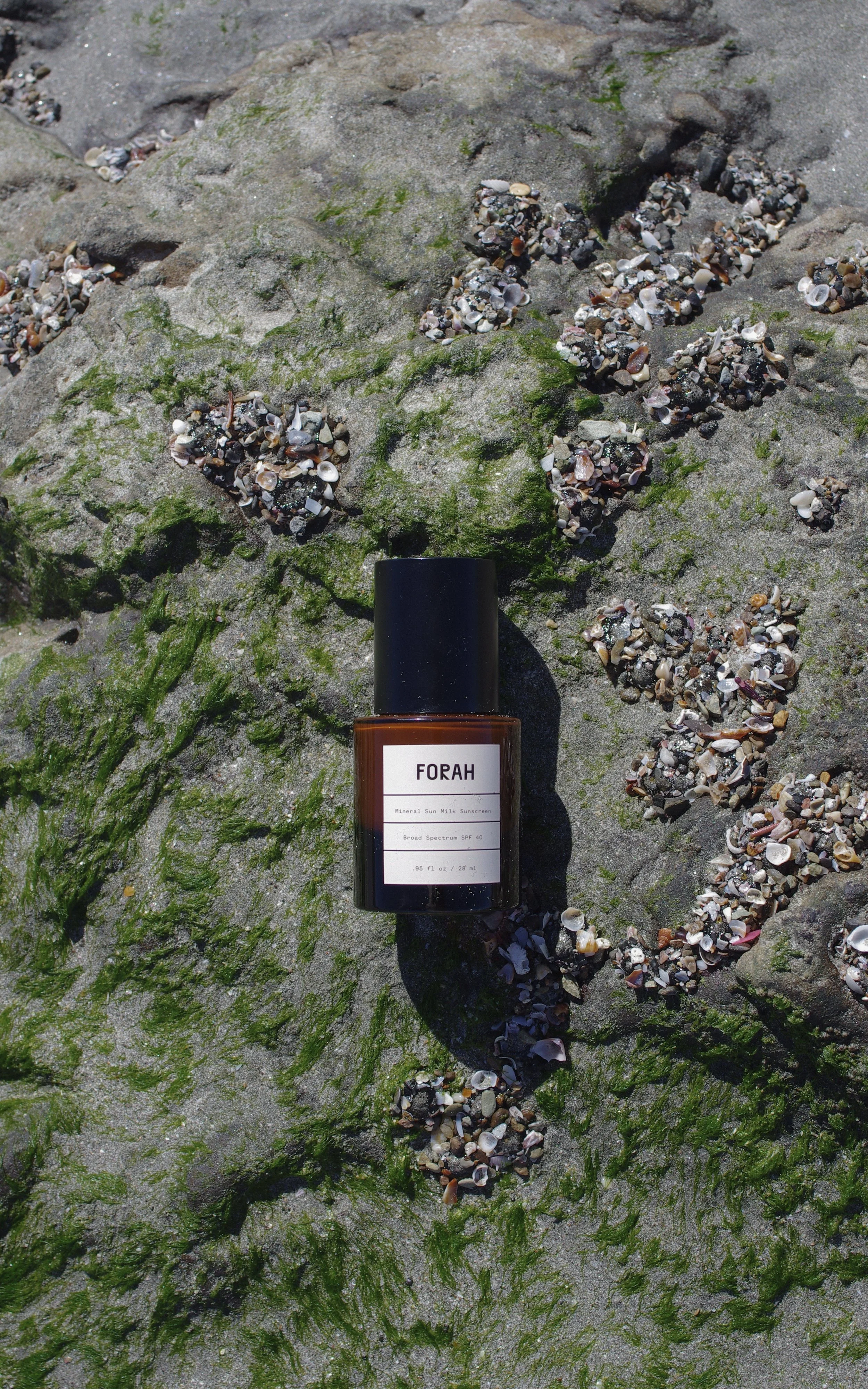 Forah Mineral Sunscreen Serum SPF40 bottle on seaweed-covered ocean rocks amid shells and moss, evoking coastal protection.