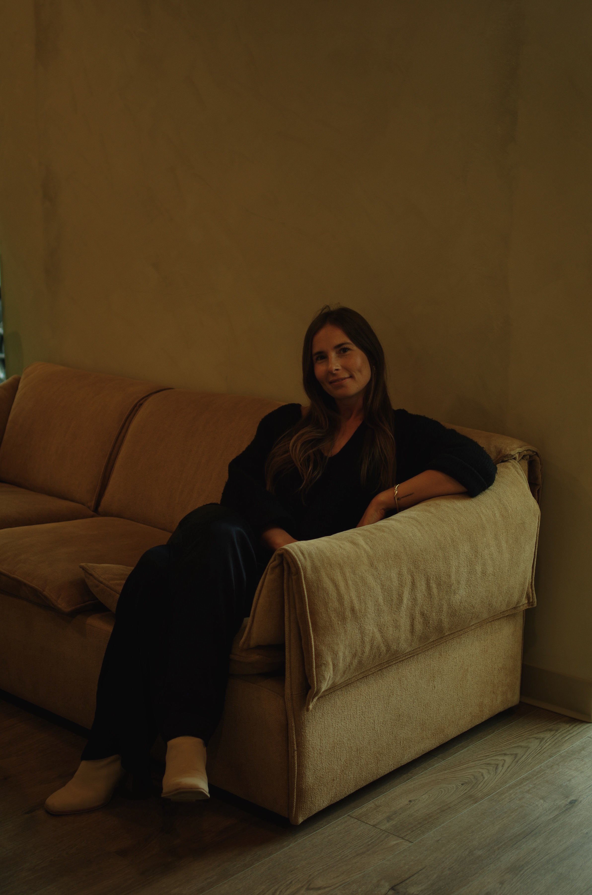 Michaela hypnotherapist lounges relaxed on a vintage tan couch in black outfit and white boots, softly smiling.