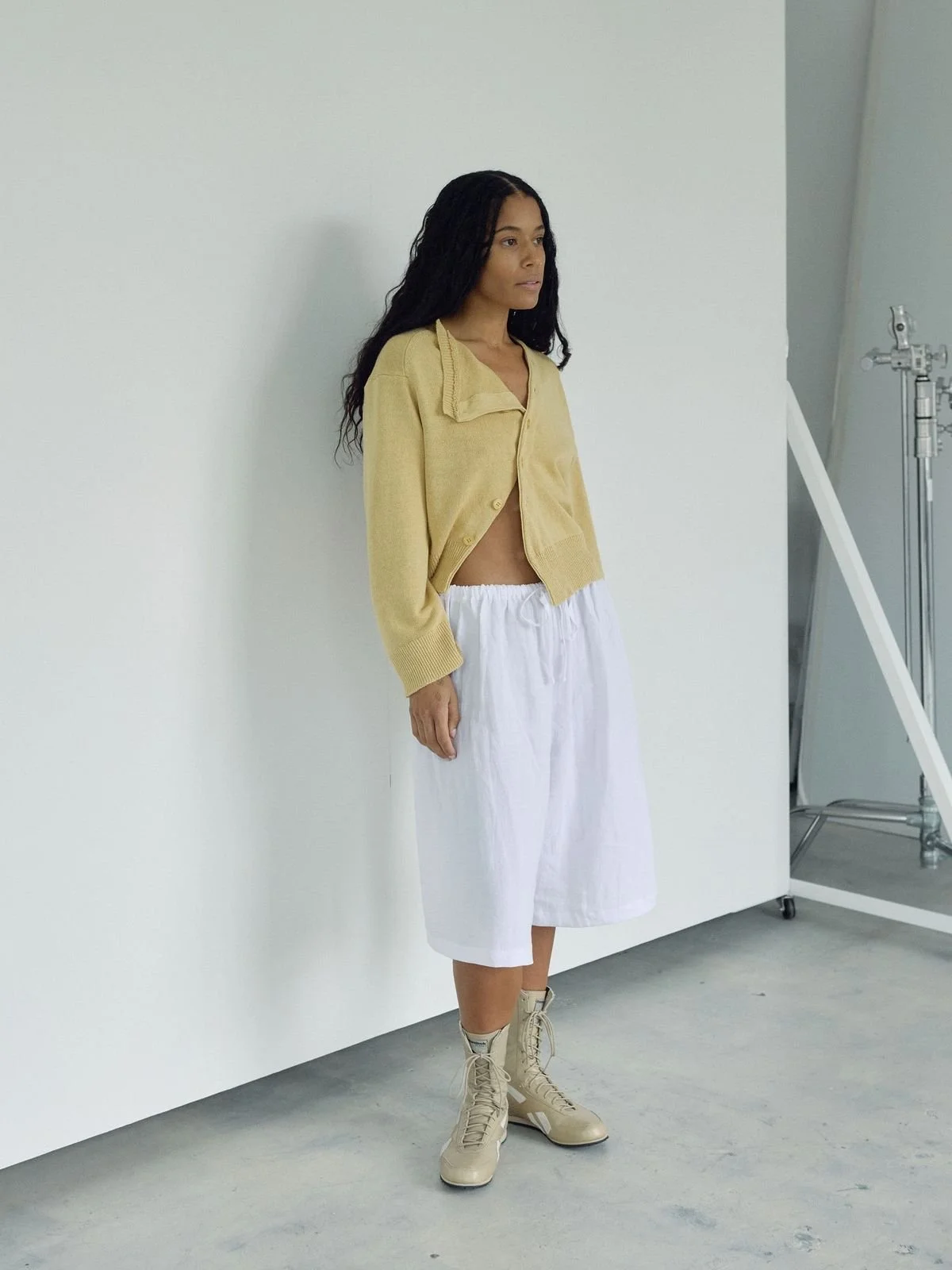 Stylish side view of model wearing Bohème Goods The Asymmetric Knit, a pale yellow open cardigan, loose white pants, and tan boots on a white studio floor.
