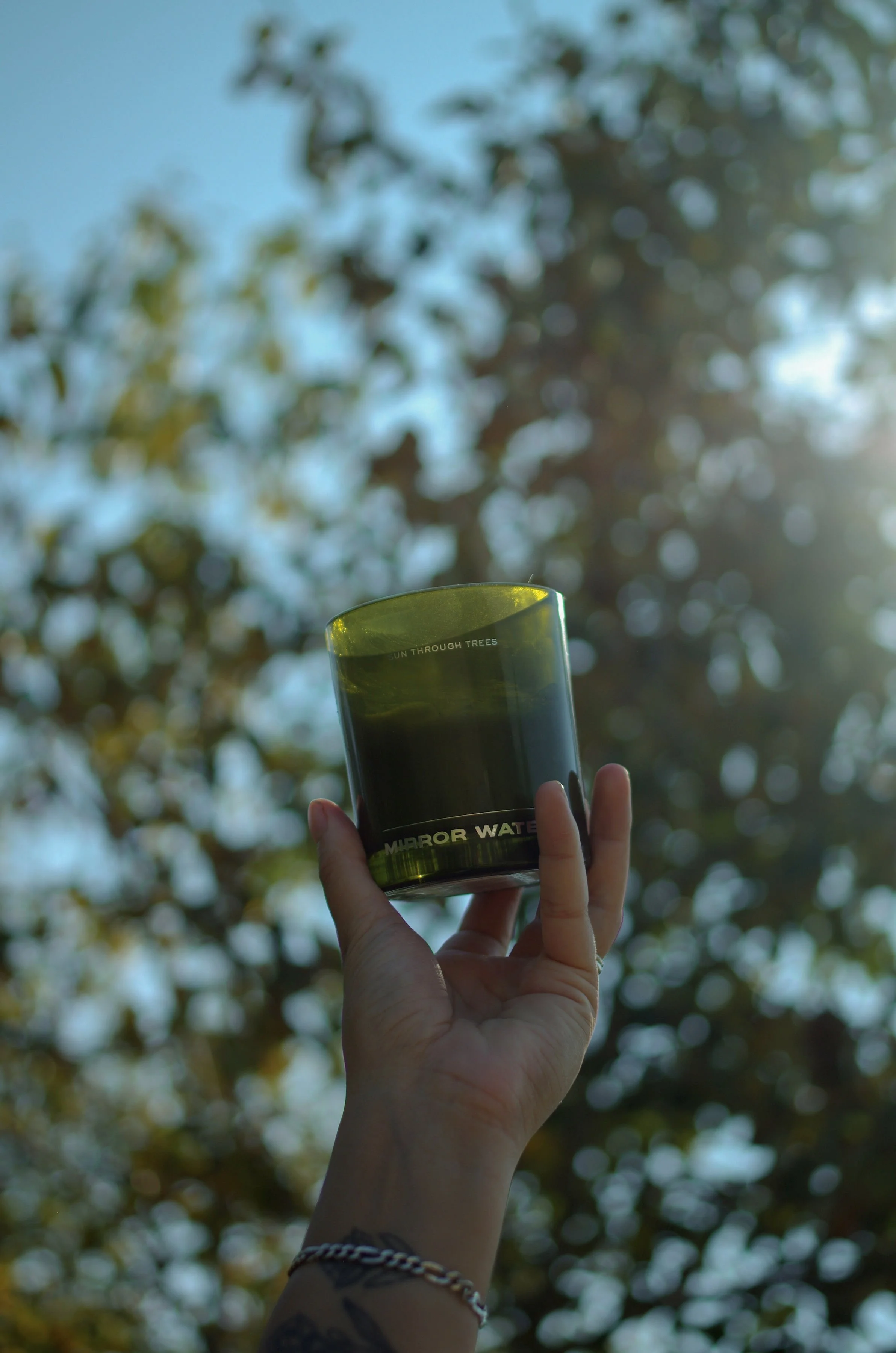 A hand holds a green glass Mirror Water Sun Through Trees Candle, with sunlight filtering through blurred trees and blue sky in Alberta.