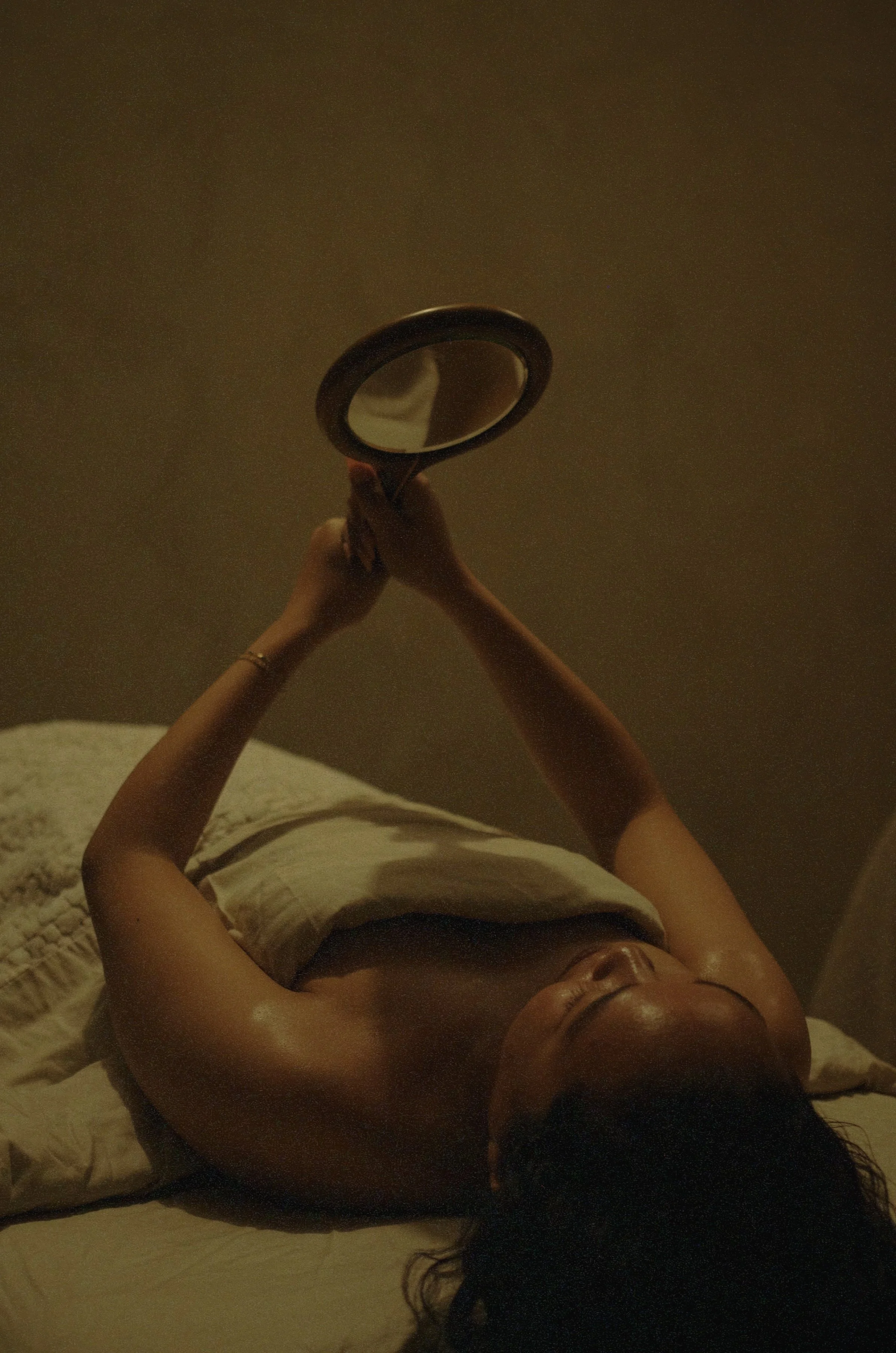 Woman with glowing skin post facial lies in bed, holding a vintage round mirror to admire her reflection.