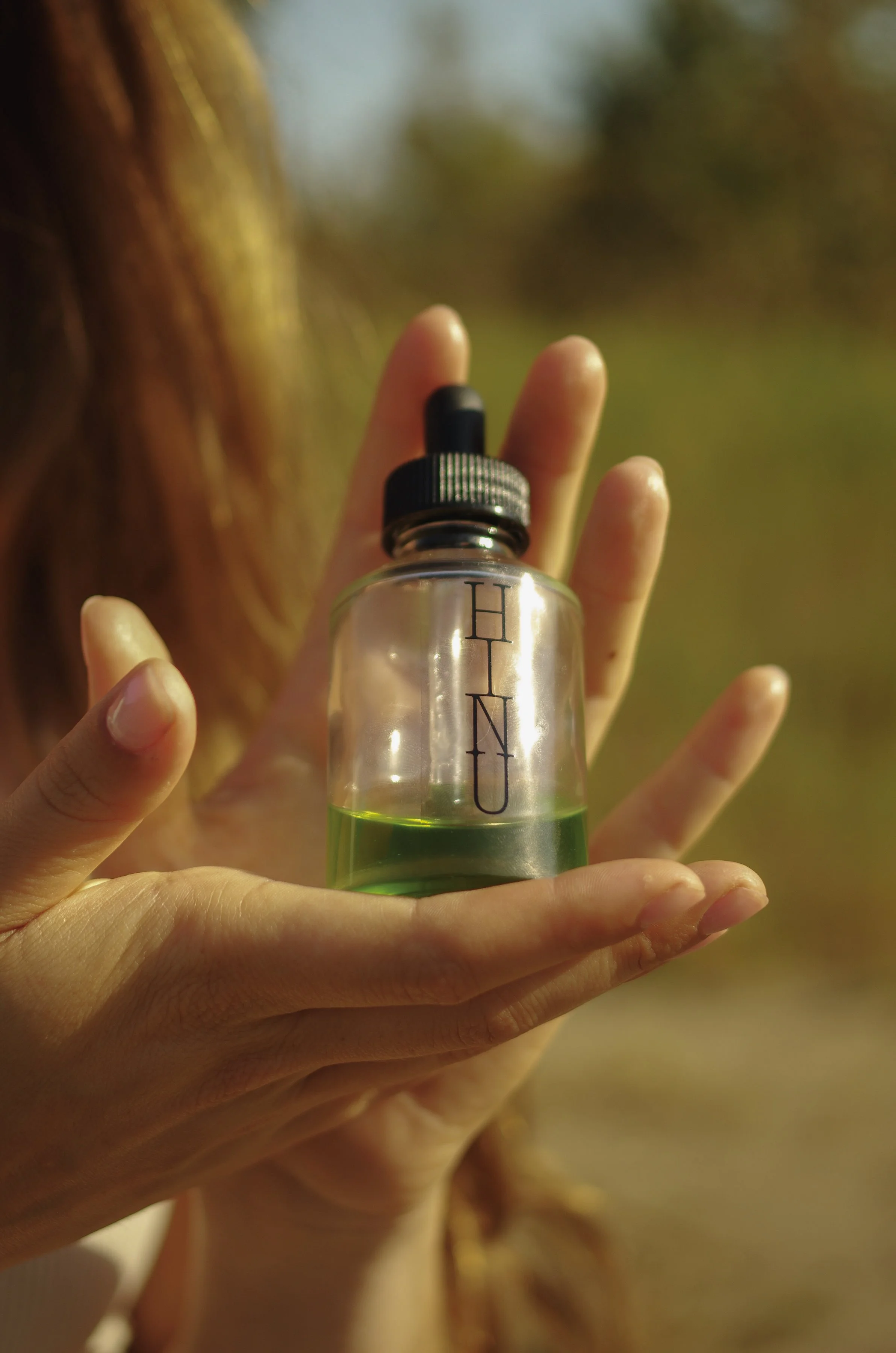Woman's hand holding a clear glass bottle of Hinu Hair Growth Oil with green liquid and black dropper cap, outdoors in green field.