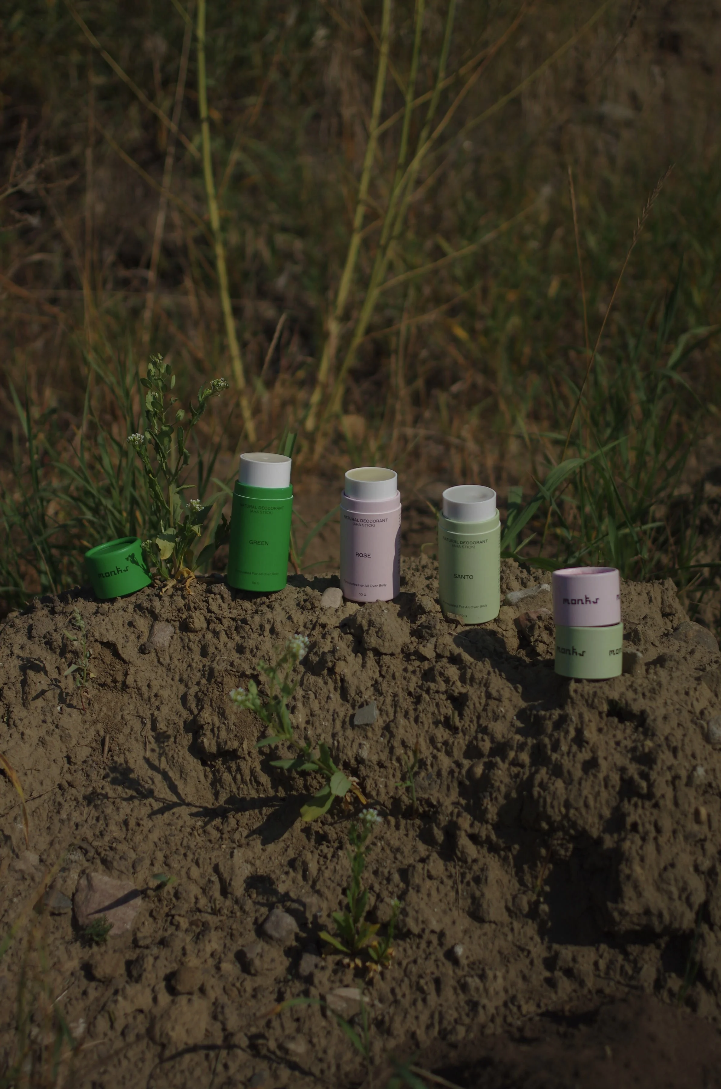Assortment of Monks Natural Deodorant Sticks in green, palo, and rose on soil with surrounding plants and grass.