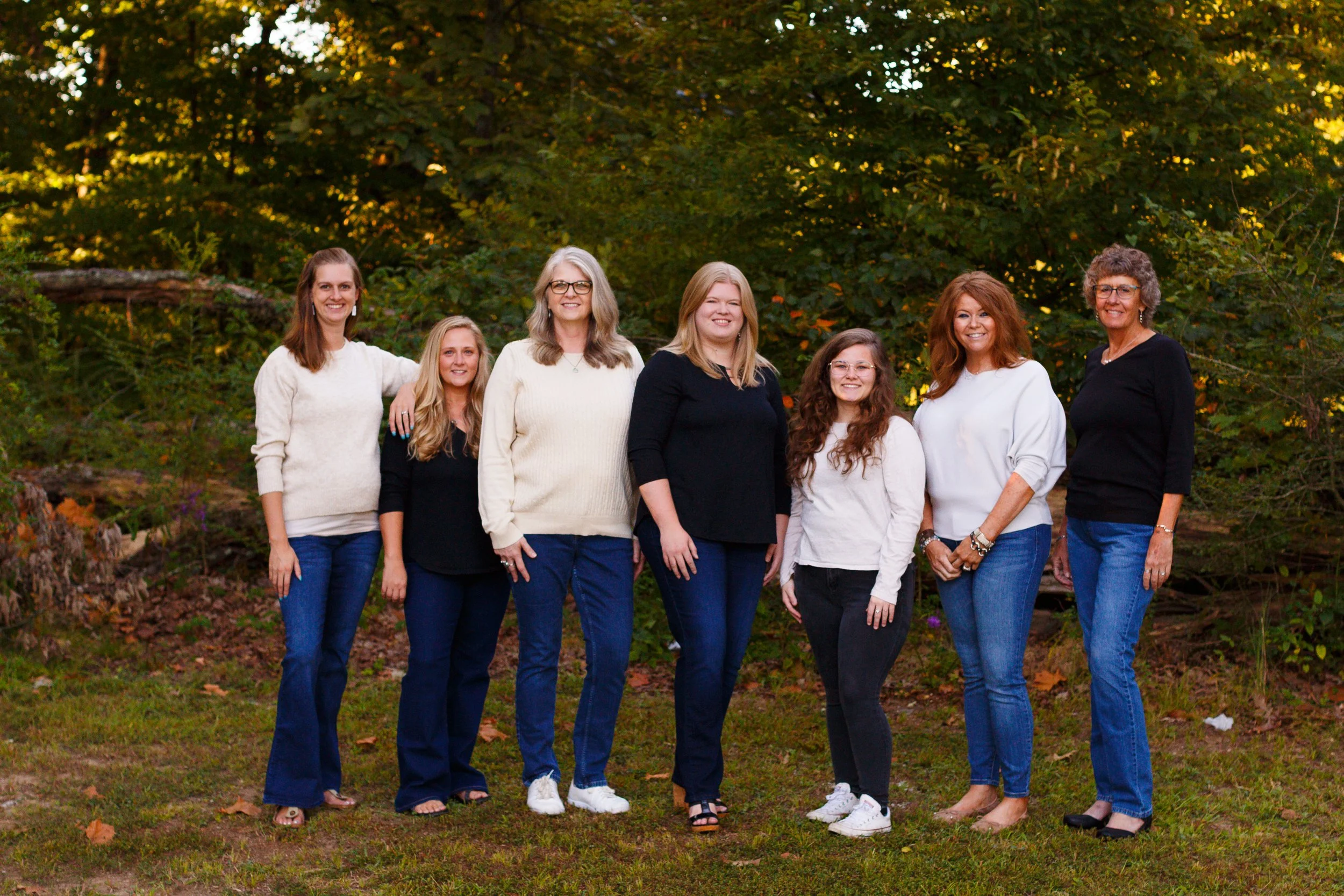 Group of seven women standing outdoors in front of trees, smiling, wearing casual fall clothing.