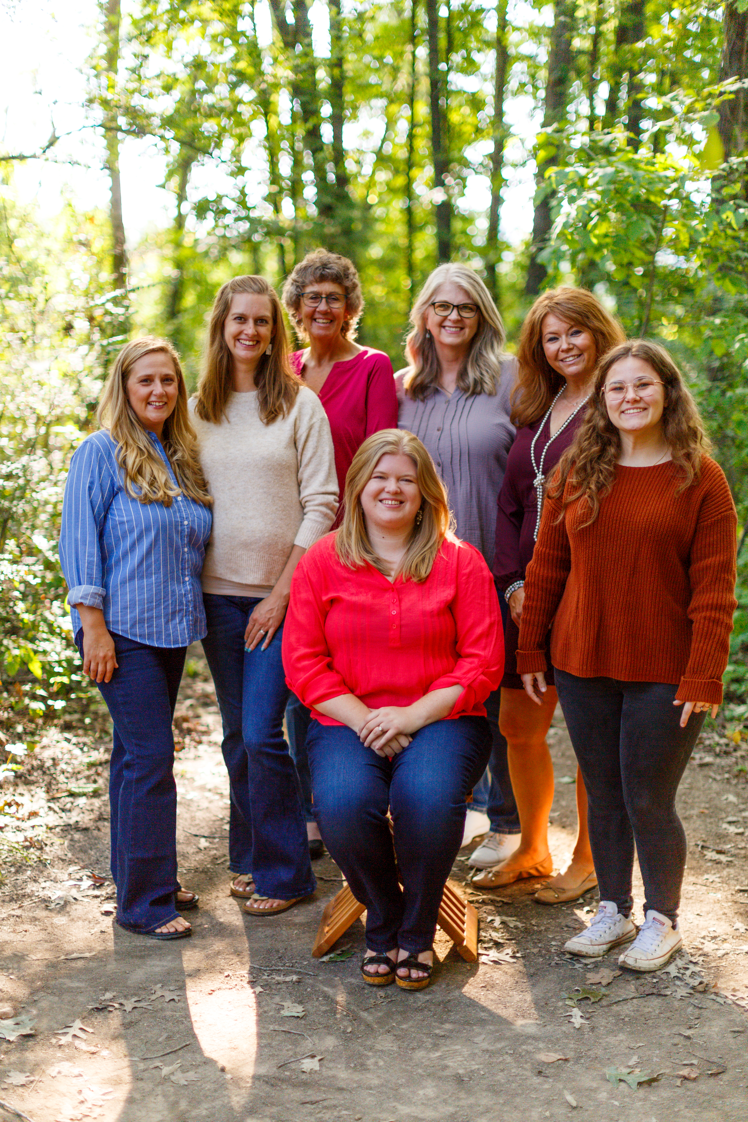 Group of eight women outdoors in a wooded area, some standing and one sitting on a small wooden stool, smiling at the camera, dressed casually in colorful tops and jeans.