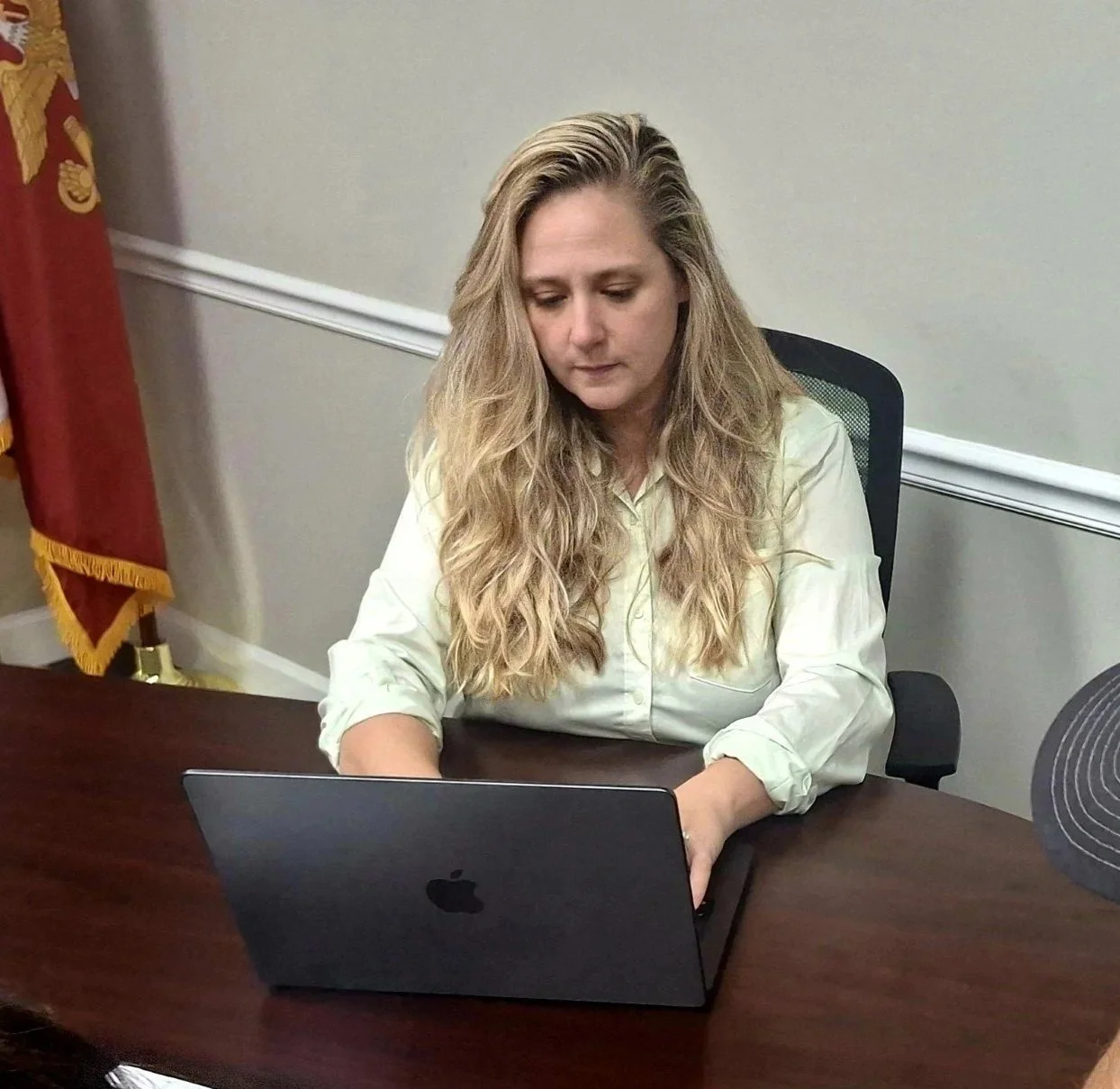 A woman with long wavy blonde hair sitting at a wooden desk, working on a silver Apple MacBook laptop in a meeting room with beige walls, white trim, and a red and gold flag in the background.