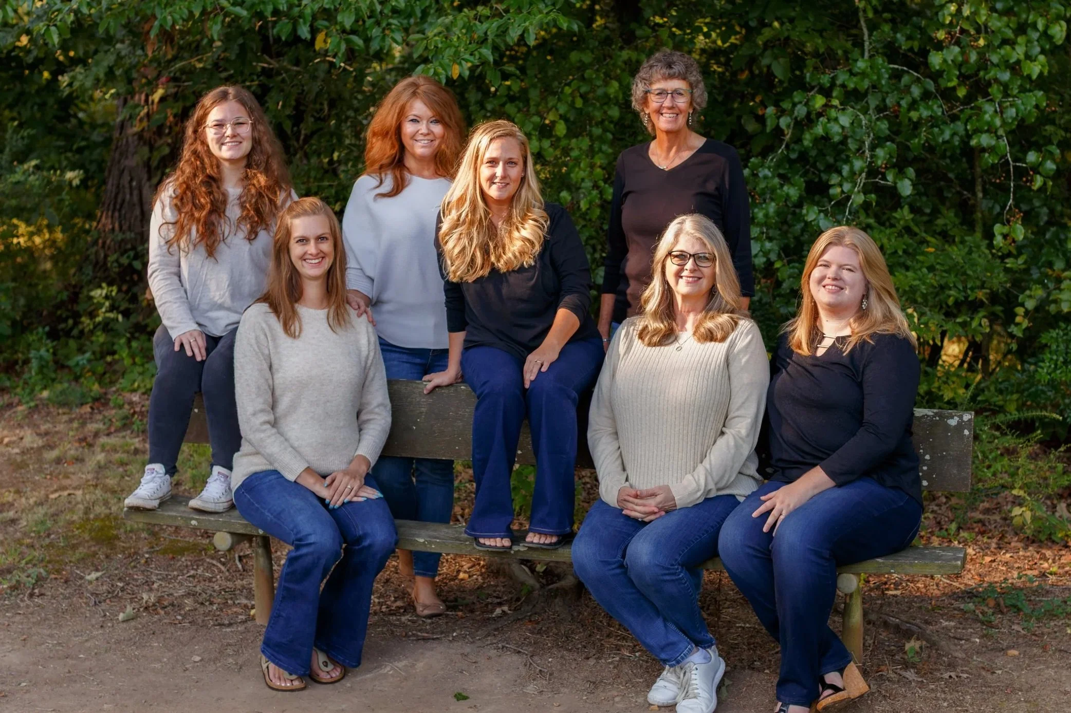 A group of nine women gathered outdoors, posing confidently for a photo in front of green trees, some seated on a wooden bench and others standing behind it.