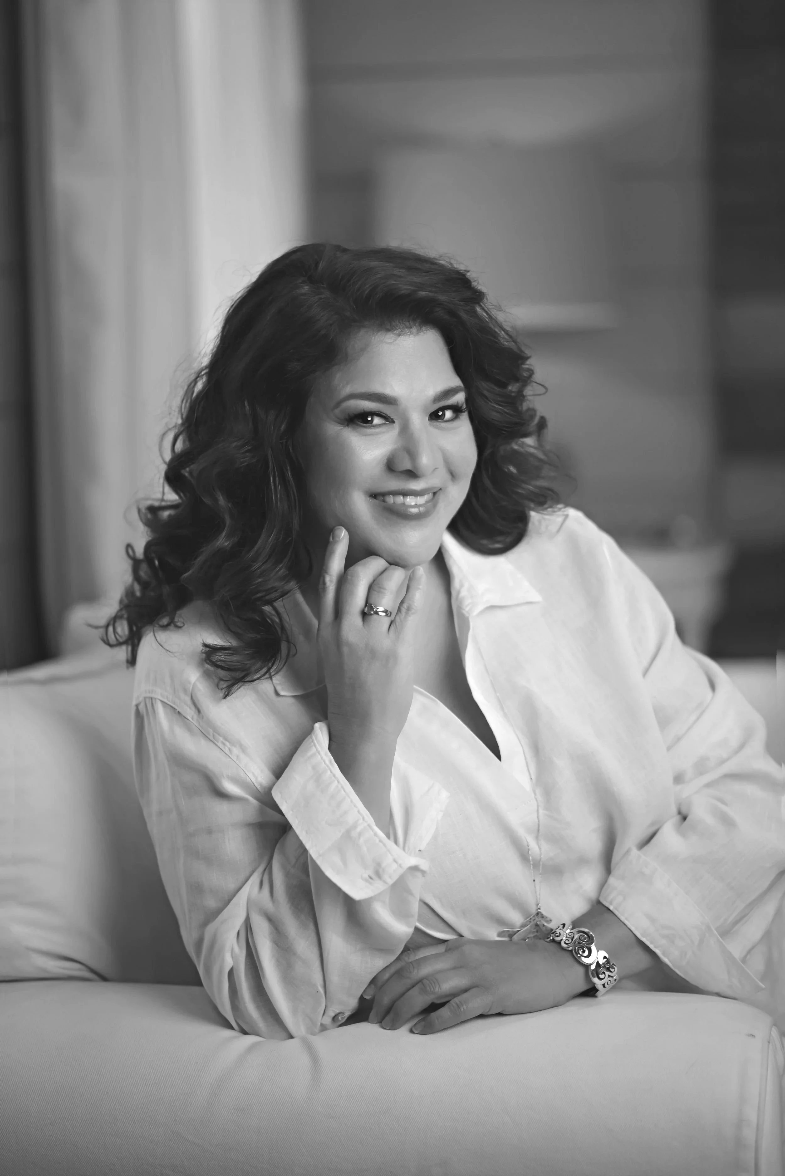Smiling woman with dark wavy hair, wearing a white blouse, sitting on a light-colored sofa in a warmly lit room.
