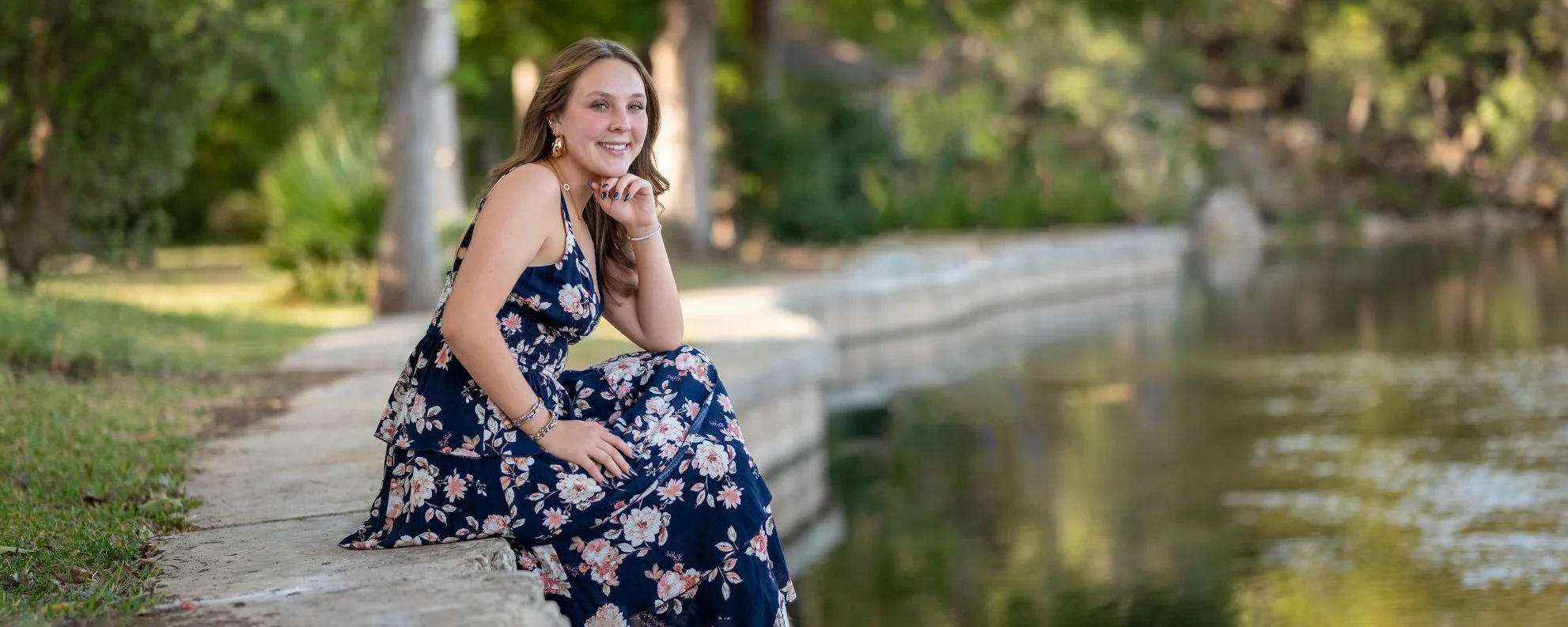 High school senior portrait at Landa Park in New Braunfels featuring a senior in a floral dress beside the water and mature trees.