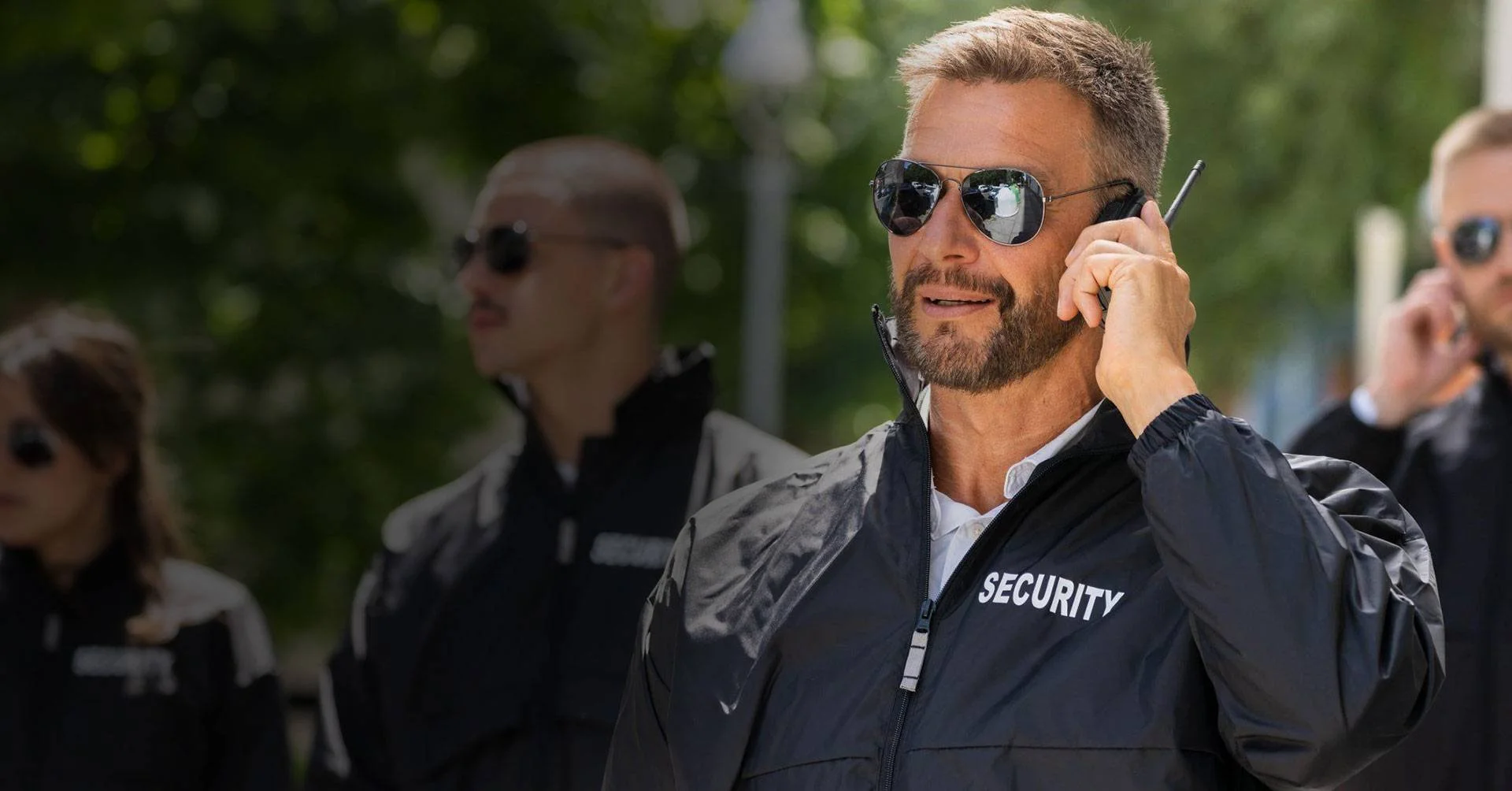 Security team leader with a beard and sunglasses using a walkie-talkie outdoors, wearing a black jacket labeled 'SECURITY,' with additional security personnel in the background—representing event and perimeter security services.