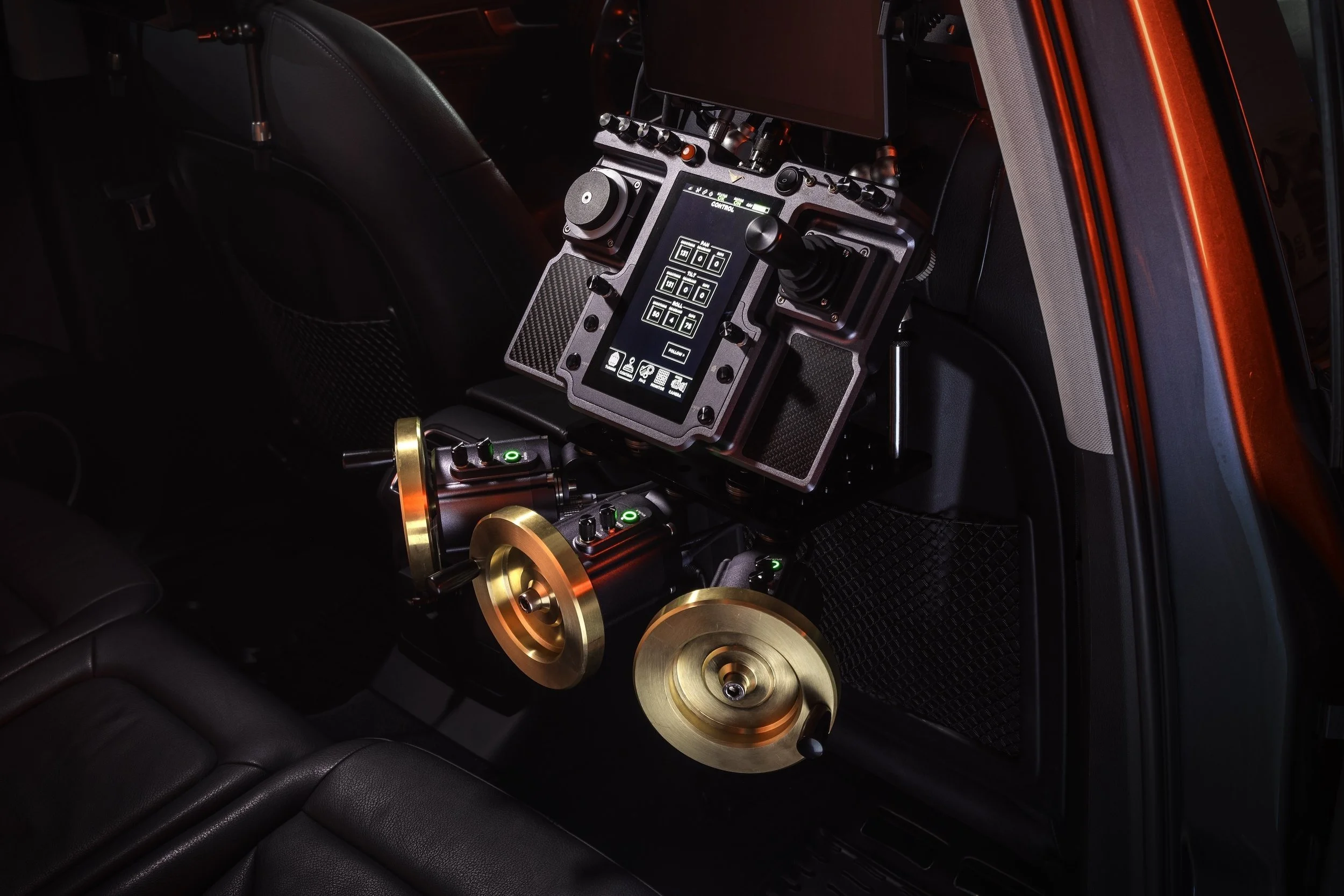 Inside a vehicle, there is a mounted control panel with a screen and physical controls, along with advanced camera or sensor equipment with large gold-colored wheels attached, positioned in the back seat area.