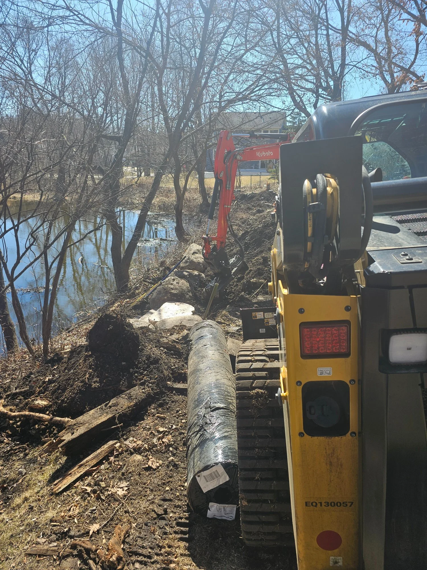 Pond boulder wall excavation with boulder fabric — Kubota and CAT on site — Woodbury MN — Heritage Outdoors