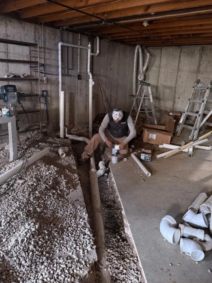 A person working on plumbing pipes in a basement or utility room, surrounded by construction tools, pipes, and ladders.