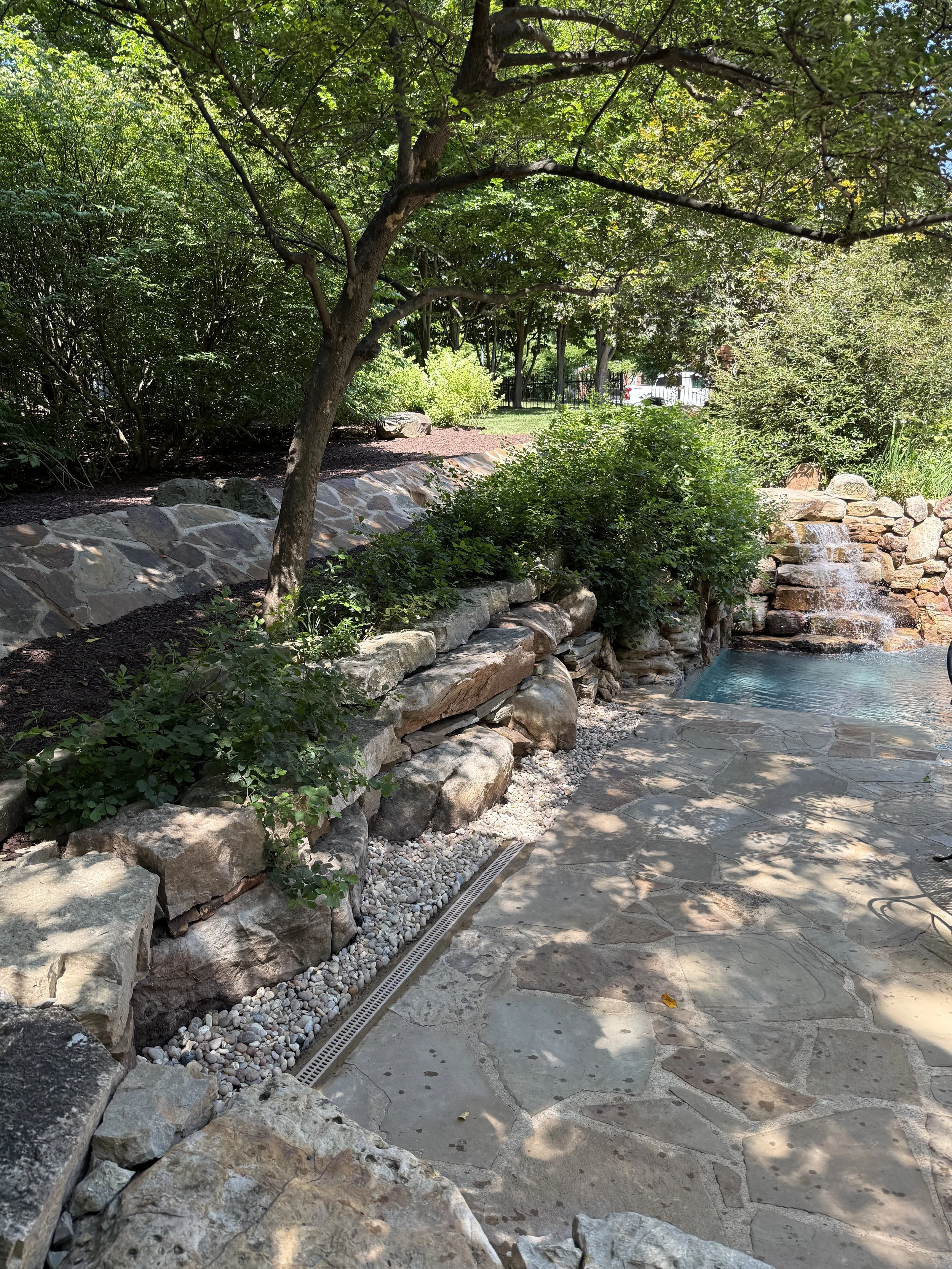 A backyard garden scene with a stone patio, a waterfall feature, lush green trees, and bushes creating a shaded, serene environment.