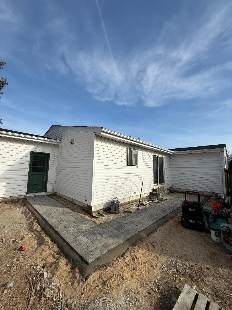 Backyard of a house under construction with newly paved walkway, construction tools, and equipment near the house, under a partly cloudy sky.