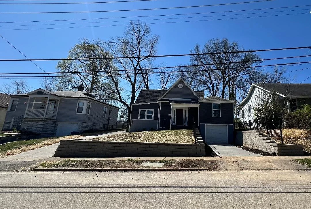 A black house with a garage on the right side, a small porch in front, and a yard with patchy grass. Two houses are on either side with clear blue sky and trees in the background.